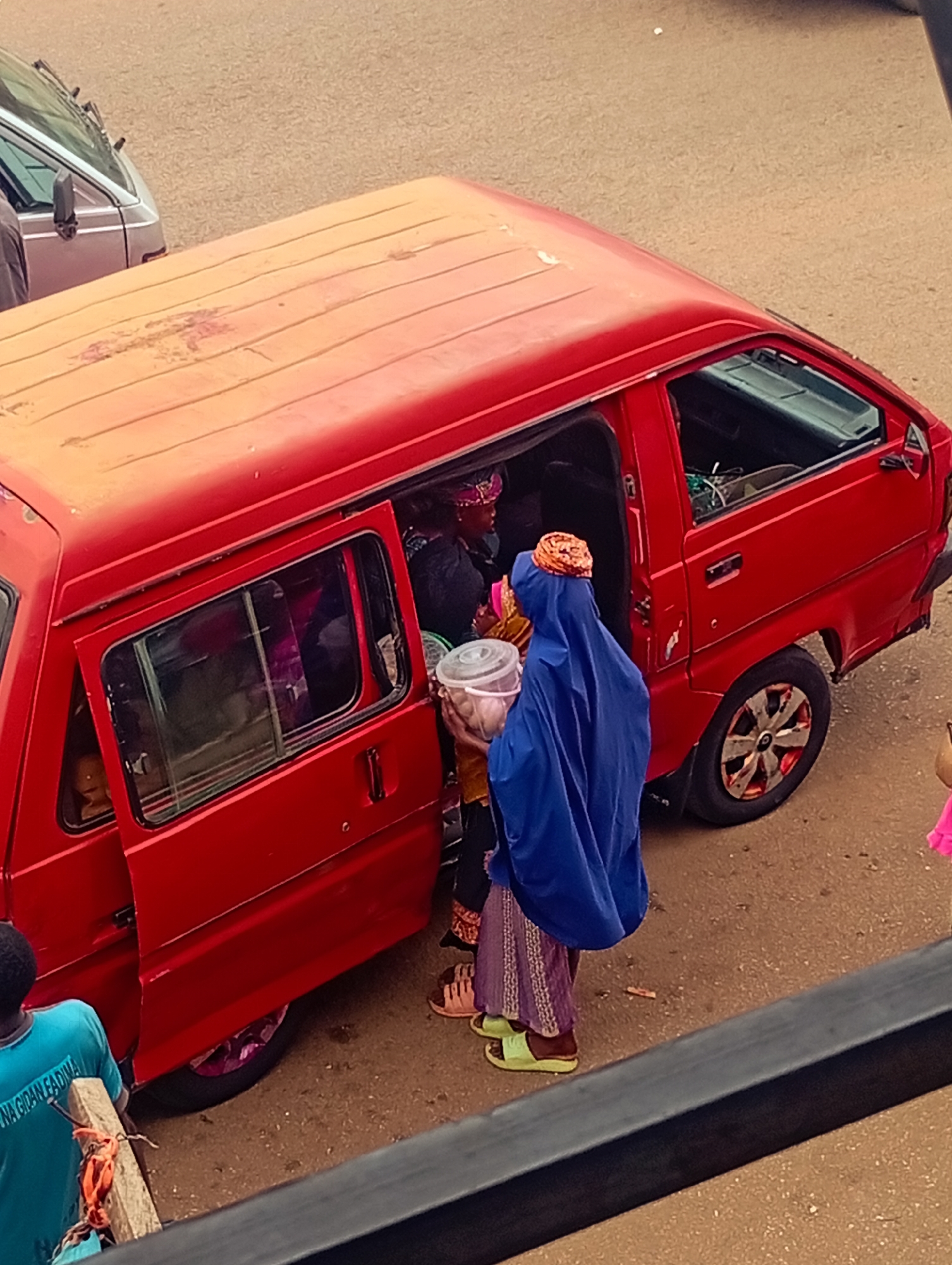 A high-angle, medium shot shows a red van with its side door open, situated on a street in an urban setting, likely in the city of Unknown, Unknown. A woman draped in a blue hijab is handing a container to a person inside the van. Other occupants are visible inside the vehicle. The ground is paved and appears to be part of a roadway. The overall lighting suggests it is daytime. Another vehicle, possibly a silver van, is visible in the background. In the lower left corner, a person wearing a blue shirt is partially visible.