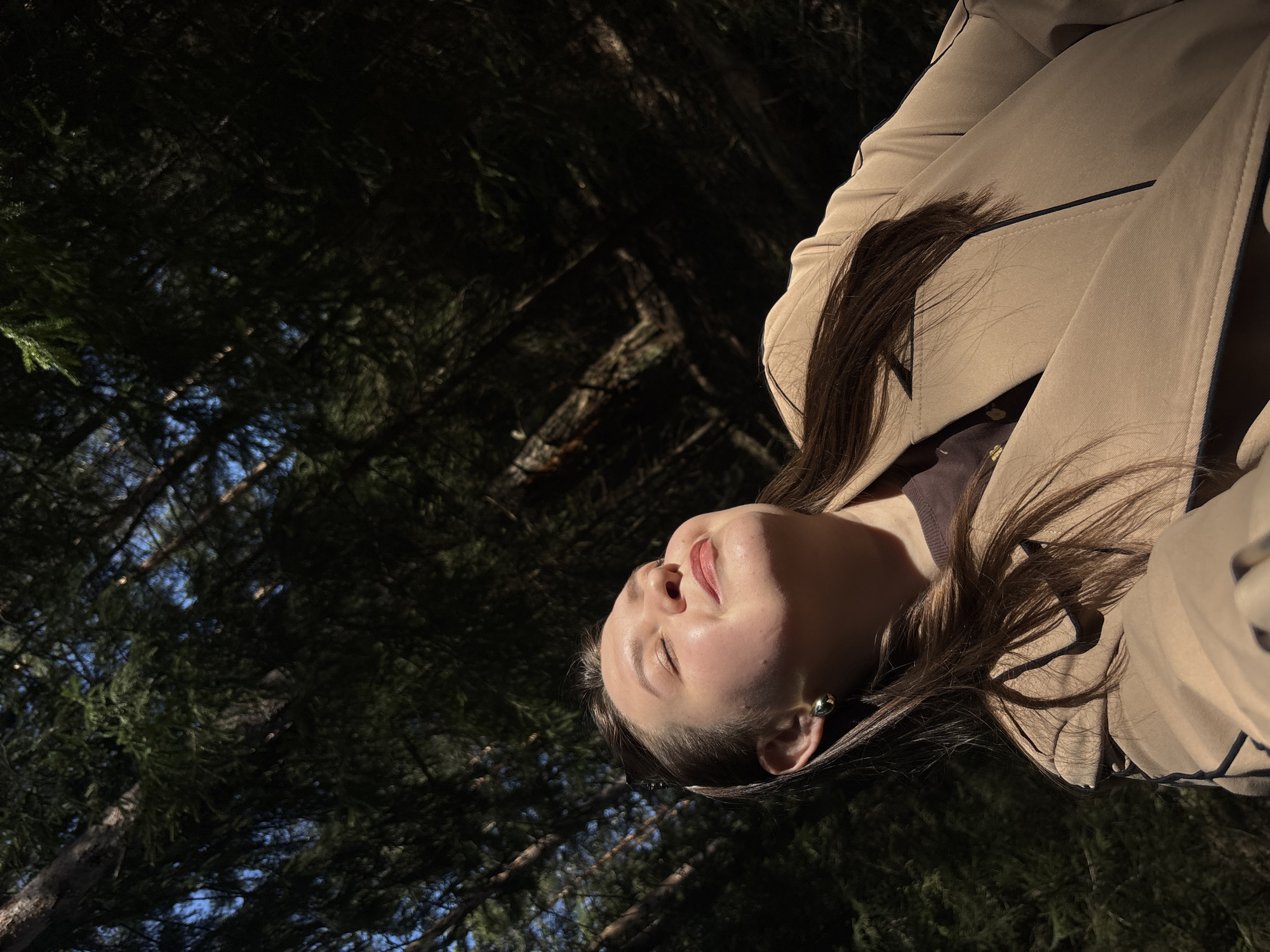 The image shows a woman in a tan trench coat lying on the ground. Her head is tilted back, face looking towards the sky. She has brown hair. Sunlight shines on her face. The background is a dense forest with pine trees. The location appears to be a forest in Марковский, Russia.
