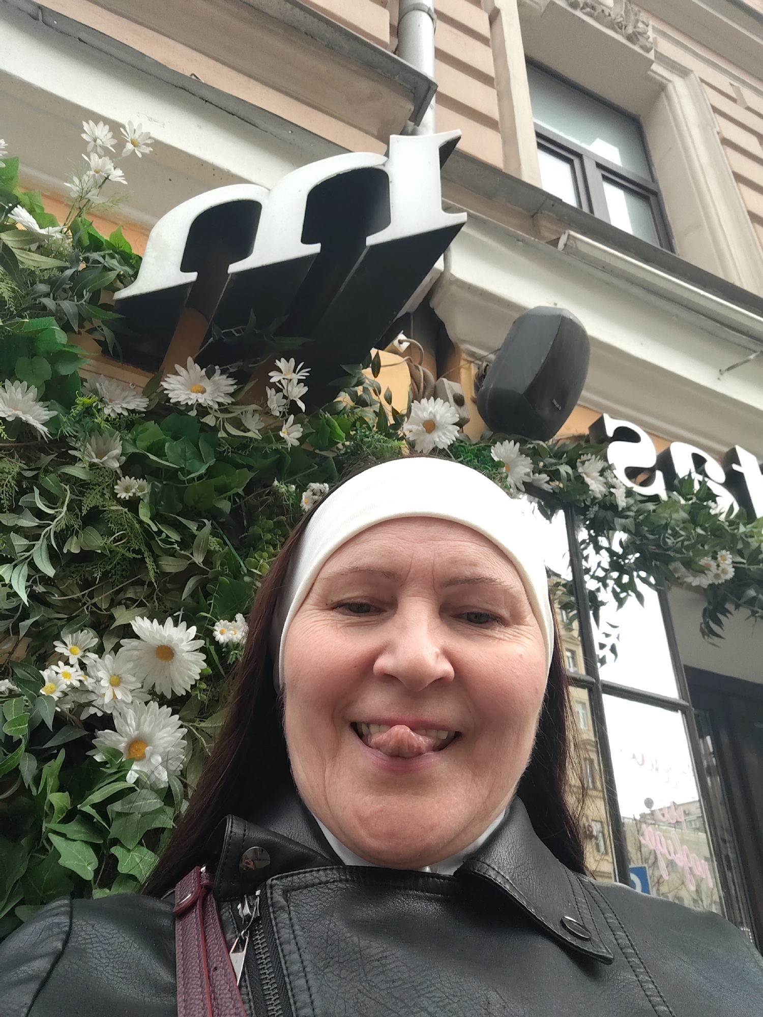 A woman in a white headband and black leather jacket is taking a selfie in front of an establishment with floral decorations and the letter "m" visible on its signage. The building appears to have windows and architectural details. The woman is sticking out her tongue. The image was taken in Moscow, Russia.