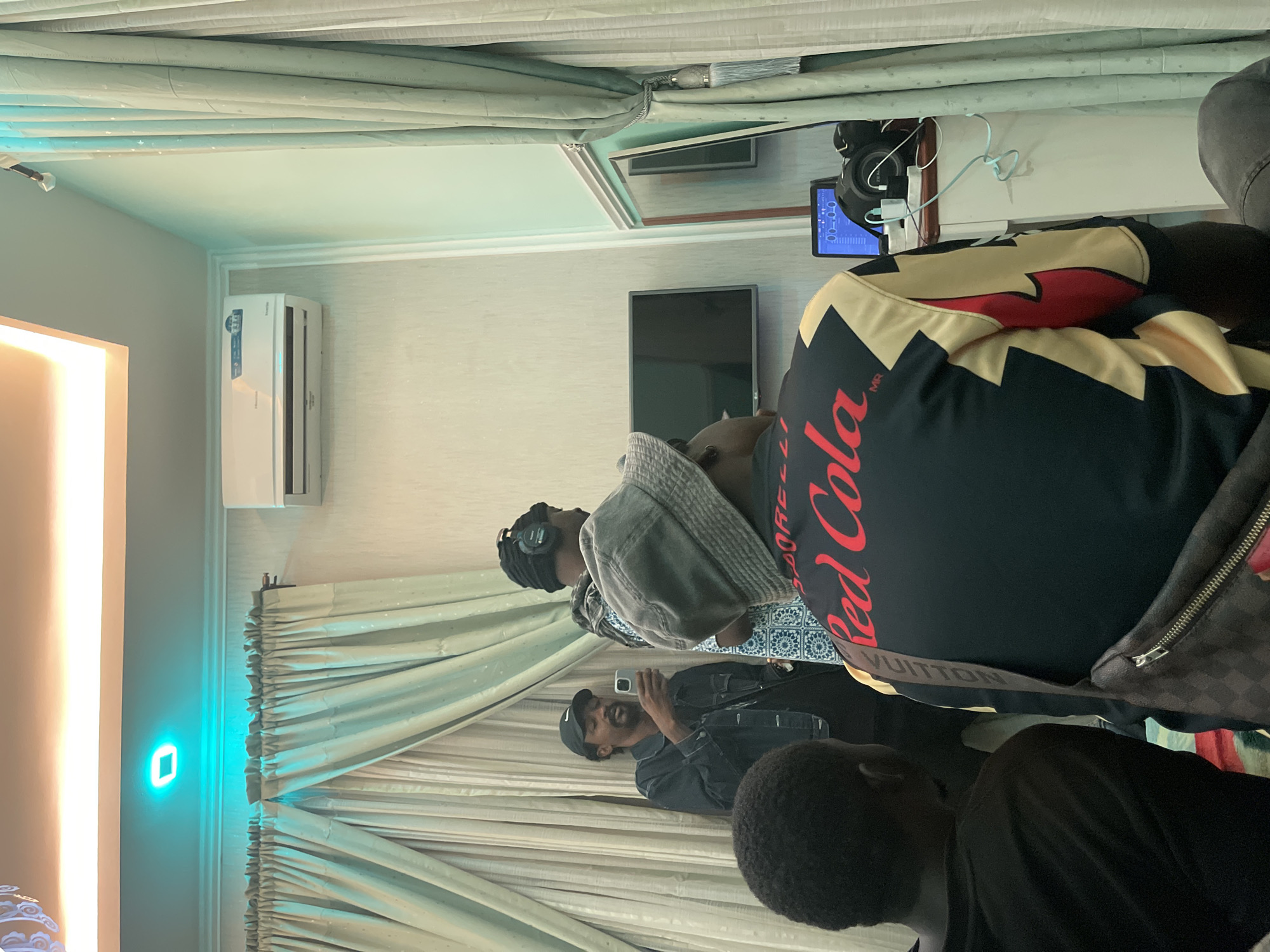 A horizontal indoor shot in what appears to be a room in Gura Top, Nigeria. Four black individuals are visible. One person wears a black shirt with "Red Cola" printed in red, a Louis Vuitton belt bag, and a jacket with black, yellow, and red patterns. Another person is wearing headphones and a gray sweater. A third person is holding a phone, possibly taking a photo, wearing a black cap and jacket. The fourth person wears a black shirt. In the background, there are patterned curtains, a wall-mounted air conditioning unit, a black flat-screen TV, and a mirror reflecting a camera set-up. A green wall light is visible on the left.