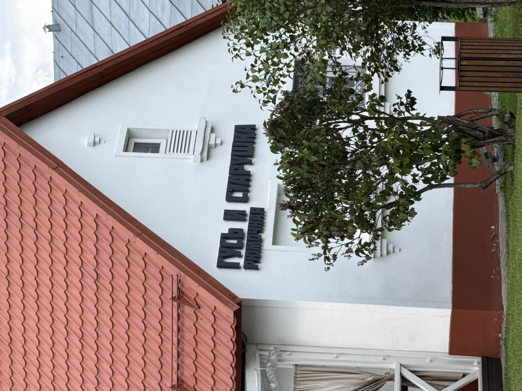 The image shows a white building with a red-tiled roof. Black lettering on the side of the building reads "ГУСЬ И СТАРУШКА" above the word "РИМОЧНАЯ". A window with horizontal slats is visible on the upper part of the wall. In front of the building, there are several trees with green leaves and dark berries. To the right of the trees, a wooden trash bin with a metal lid is positioned on the grass. The scene appears to be an outdoor setting, possibly a restaurant or shop, in Moscow, Russia, indicated by the Cyrillic text. The sky is overcast.