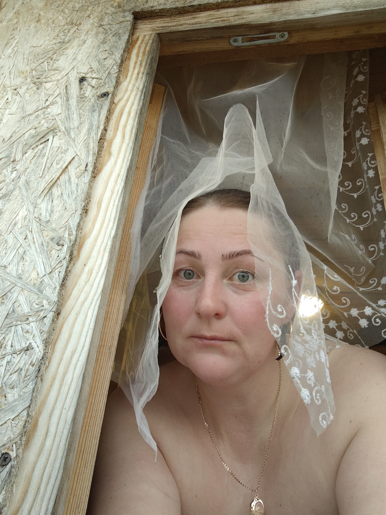 A medium, eye-level shot depicts a fair-skinned woman peering through a wooden frame, possibly a window. The woman has light blue eyes, fair eyebrows, and a slightly furrowed brow, giving her a somewhat pensive expression. Part of a sheer white lace curtain is draped over her head and shoulder. She is wearing a gold necklace with a pendant, and a gold hoop earring is visible. Her shoulders are bare, suggesting she is unclothed from the chest up. The wooden frame appears to be constructed from raw lumber, with the texture of the wood grain clearly visible. The location of Фоки, Russia cannot be confirmed based on the provided visual information.