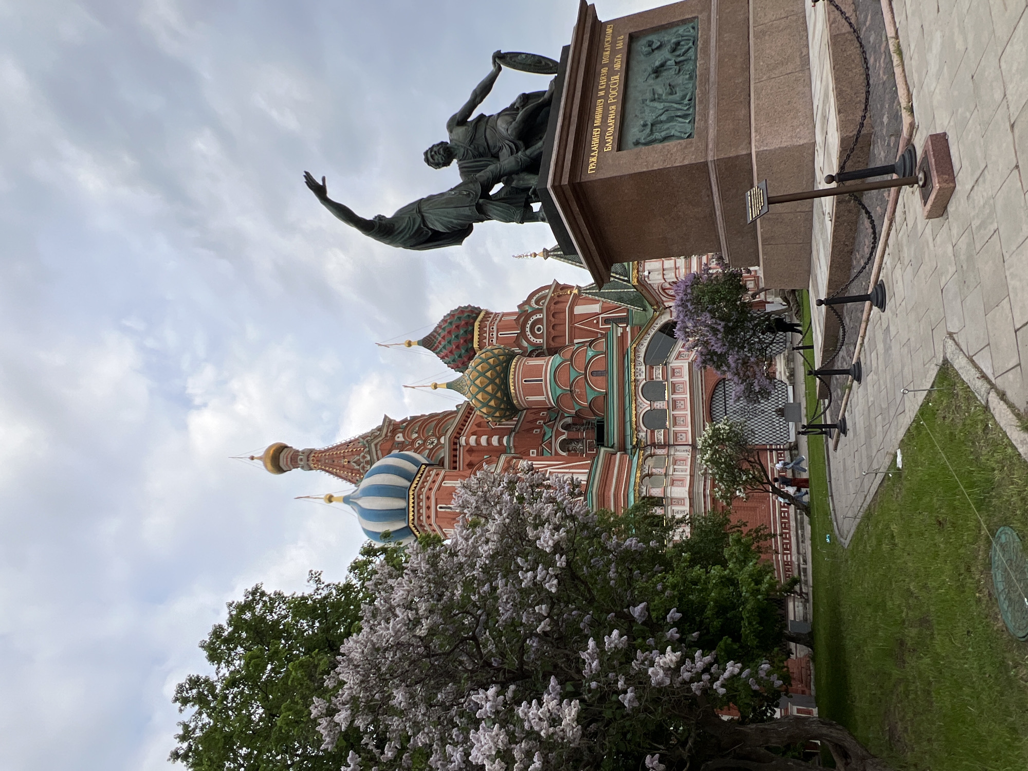 The image shows St. Basil's Cathedral in Moscow, Russia, with a monument to Minin and Pozharsky in the foreground. The cathedral features colorful domes and ornate brickwork, partially obscured by trees with white and green foliage. The bronze statue of Minin and Pozharsky stands on a red granite pedestal with inscriptions and reliefs. The sky is overcast. A paved walkway and grassy area are in the foreground. Several people are near the cathedral, with a man wearing blue clothing, and people with backpacks.