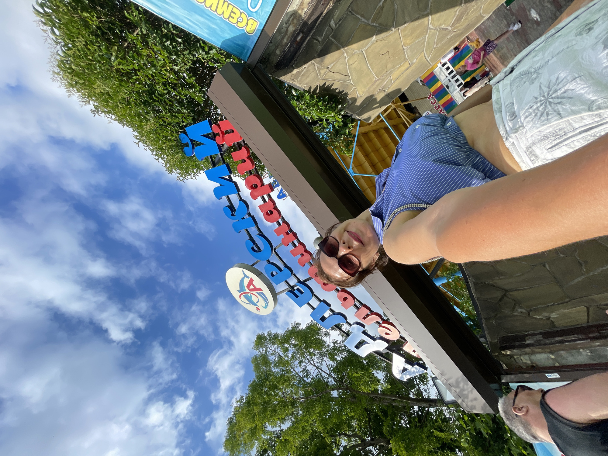 This is a low-angle, outdoor, selfie-style shot taken during the day, likely late morning or early afternoon given the bright sunlight. The subject, a woman with dark hair, sunglasses, and a blue and white striped top, is smiling at the camera. She appears to be at an amusement park or similar recreational area.
Behind her, a large sign with colorful, raised letters spells out "SOCHI PARK" in Russian. The letters are in blue, red, and white. To the left of the sign is a circular emblem with a stylized bird and a red, white, and blue design. The background shows lush green trees and a bright blue sky with scattered white clouds. The ground is paved with light-colored stones.
In the far background, there's a glimpse of a colorful structure that looks like a carousel or a small ride, with people visible on it. The overall mood is cheerful and fun, suggesting a vacation or outing. The lighting indicates it's a sunny day. The presence of the "Sochi Park" sign clearly places the location as Sochi, Russia. A man in a black tank top is partially visible in the lower right corner of the frame, also looking away from the camera.