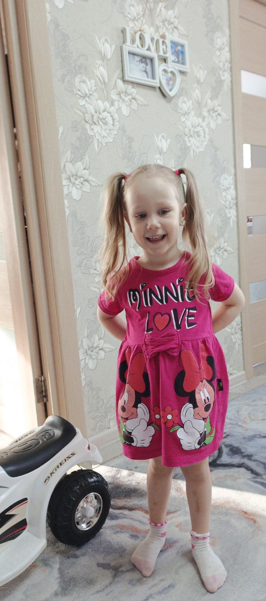 A young girl stands in a room with floral wallpaper in Chaikovsky, Russia. She wears a bright pink dress with Minnie Mouse imagery and has blonde hair in pigtails. She is smiling and has her hands on her hips. Beside her is a small, white toy vehicle with the brand "KREISS" visible. A framed decoration with the word "LOVE" hangs on the wall in the background. The floor is covered with a light-colored carpet with a swirling pattern. A door frame and curtain are partially visible on the left.