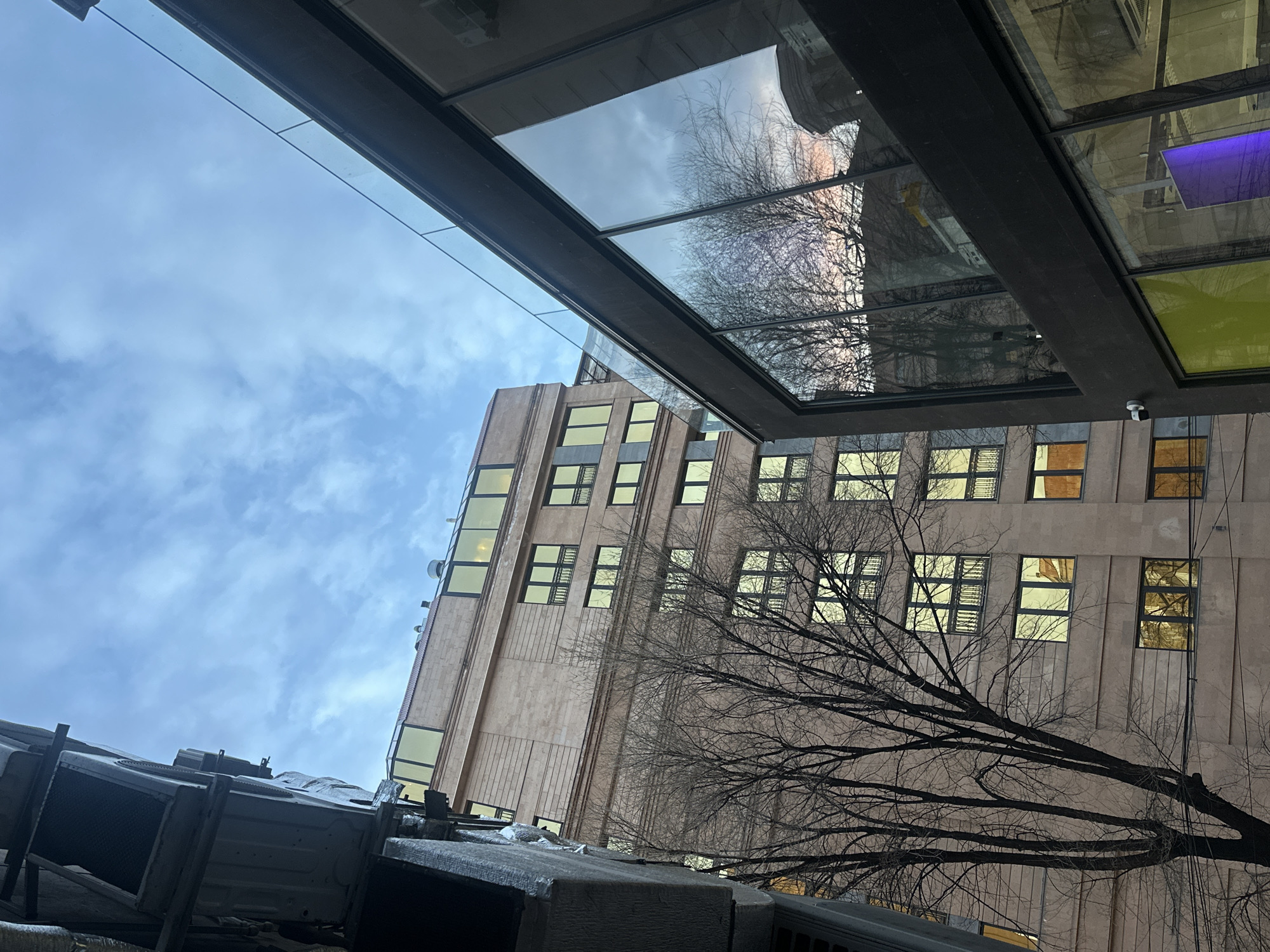 The image presents a low-angle view of a building with numerous windows, under a cloudy sky. A tree, devoid of leaves, partially obscures the building's facade. The building's windows appear to be illuminated from within. An architectural structure, possibly a glass awning, is visible in the upper portion of the frame, reflecting the sky and elements of the surrounding environment. In the foreground, sections of mechanical equipment, likely related to building systems, are visible. The exact location within Yerevan, Armenia, cannot be determined based solely on the visual information provided.