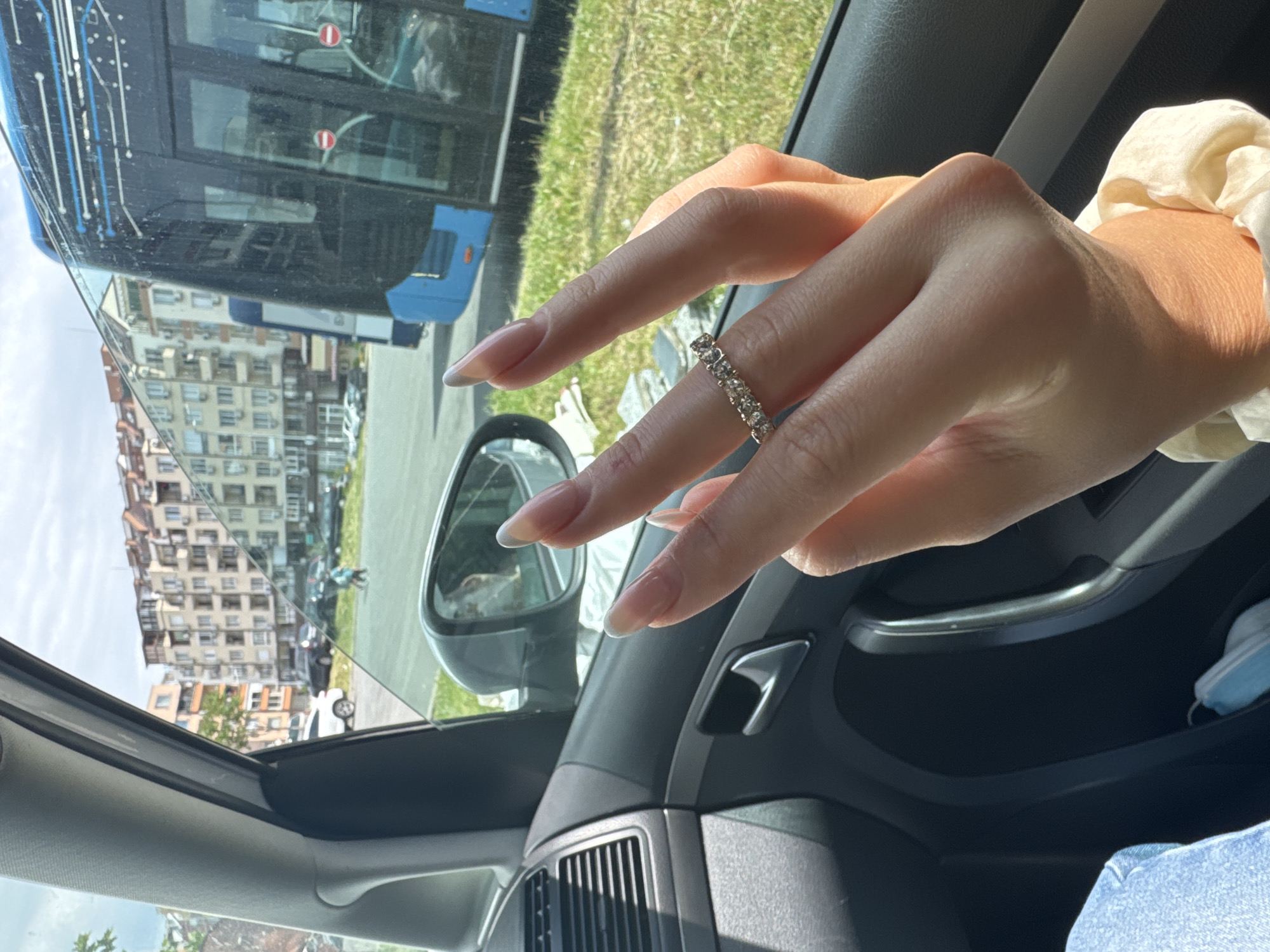 A person's hand is shown wearing a diamond ring and with long, manicured nails. The hand is positioned over the interior door of a vehicle, with the door handle and air vent visible. Outside the vehicle window, there is a reflection of a large blue bus, a multi-story building, and greenery. Based on the style of architecture and the specific bus, the location is likely in Burgas, Bulgaria. The light suggests it is daytime. A light blue surgical mask is visible in the car door pocket. The person is wearing a light blue pair of jeans and a silk looking scrunchie.