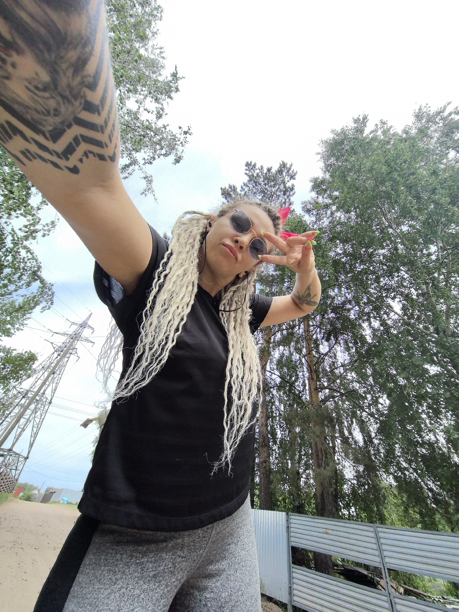 A woman is taking a selfie outdoors in Noviy, Russia. She is wearing sunglasses, a black t-shirt, and gray sweatpants. She has blonde dreadlocks and a tattoo on her arm, which is extended towards the camera. She is making a peace sign with her other hand. In the background, there are trees, power lines, a dirt road, and a fence. The sky is overcast.