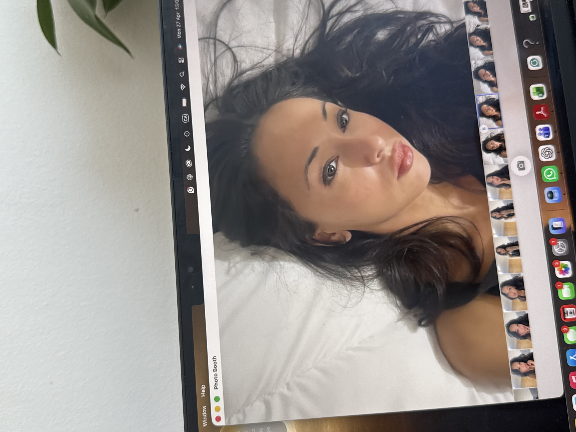 The media file captures a close-up, slightly angled shot of a laptop screen displaying a Photo Booth session. The screen is oriented vertically, showing a young woman with long, dark hair and a dark top, lying on a white surface, likely a pillow or bed. She has a posed expression, looking towards the camera with slightly parted lips.
On the laptop screen, to the right of the main image, a column of smaller thumbnail images shows multiple similar self-portraits of the woman, indicating she was taking a series of photos. The macOS interface is visible, with "Photo Booth" and "Window Help" at the top left of the application window. The top right corner of the macOS menu bar shows the date and time: "Mon 27 Apr 13:00". Various application icons are visible in the dock on the right side of the screen, including search, Wi-Fi, battery, and several common communication and system applications, some with notification badges (e.g., "3" on a settings-like icon).
A small part of a green plant is visible in the upper left corner of the physical image, outside the laptop screen. The laptop screen itself exhibits some glare, particularly on the left side. The scene is set indoors, reflecting a personal moment of self-photography, likely in a residence in London, United Kingdom.