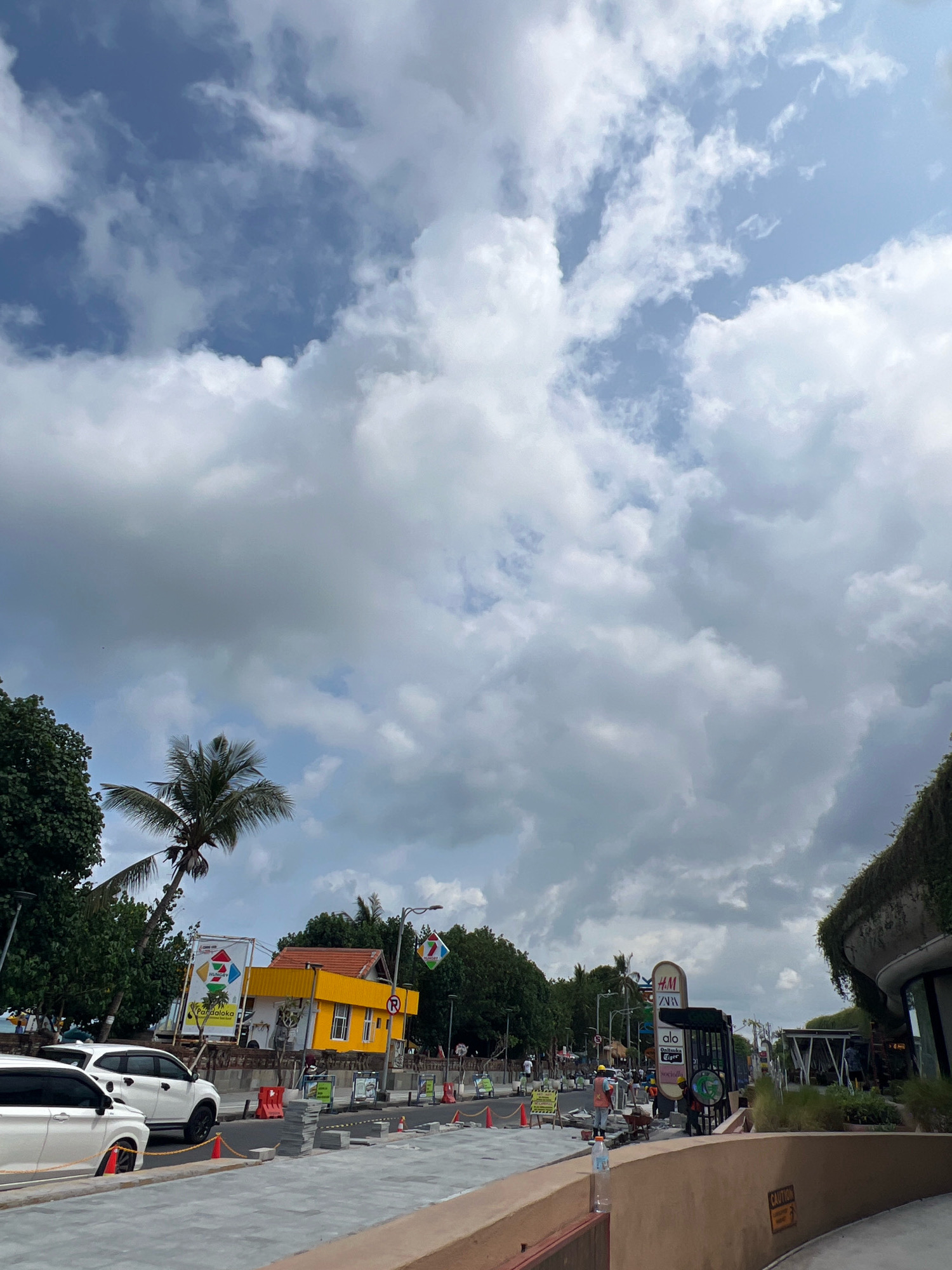 The image depicts a street scene in Kuta, Indonesia, under a cloudy sky. In the foreground, a tan-colored wall curves to the right, with a "CAUTION" sign visible on its side. The street is paved with gray tiles, and construction materials, including stacks of gray bricks and orange traffic cones, are present. Several white vehicles are parked along the left side of the street.
Further down the road, a bright yellow building with a red roof stands out, adorned with signage, including one that reads "PARADOKA." To the left of the yellow building, a palm tree is visible. In the distance, trees line the street, and there are signs for businesses such as H&M and Zara. Several people are present, some appearing to be workers engaged in construction, with one individual pushing a wheelbarrow. The scene suggests an active urban environment with ongoing development or maintenance.