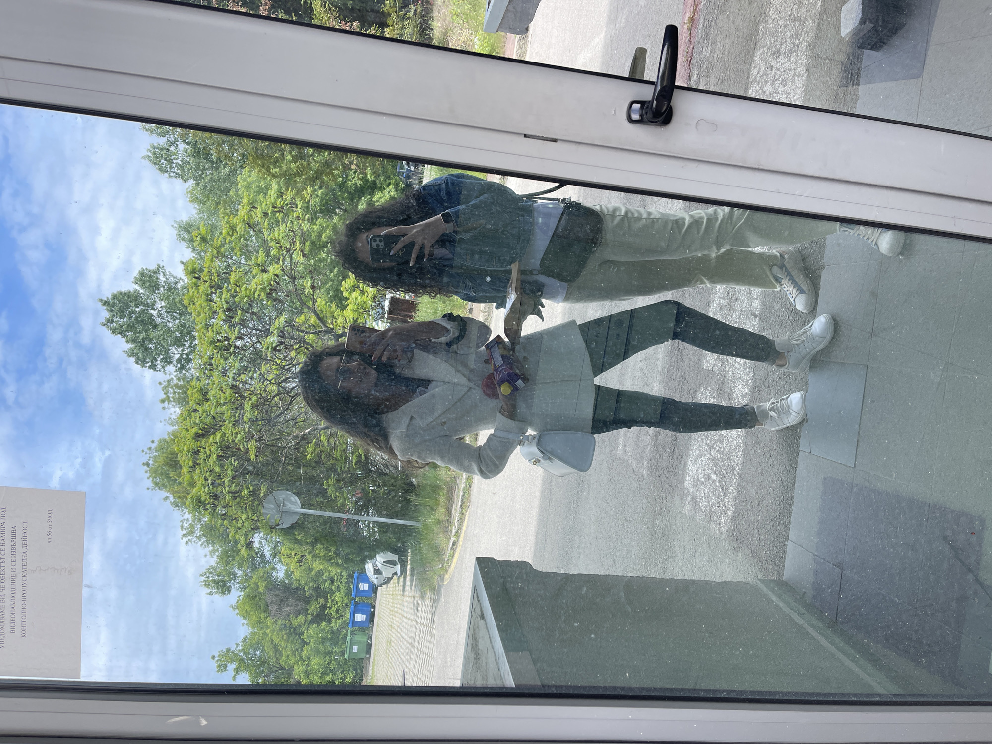 The image shows a reflection in a window, likely in Sofia, Bulgaria, based on the Cyrillic text on a sign visible in the reflection. Two women are reflected in the window. The woman closer to the window is wearing a light-colored blazer, dark pants, and white sneakers. She is holding a light-colored purse and what appears to be a bag of sweets. The other woman is wearing a denim jacket, light-colored pants, and white sneakers. She is holding a cell phone and appears to be taking a picture. In the background, visible through the reflection, are trees, a parking lot, and blue and green recycling bins. The day appears to be sunny.
