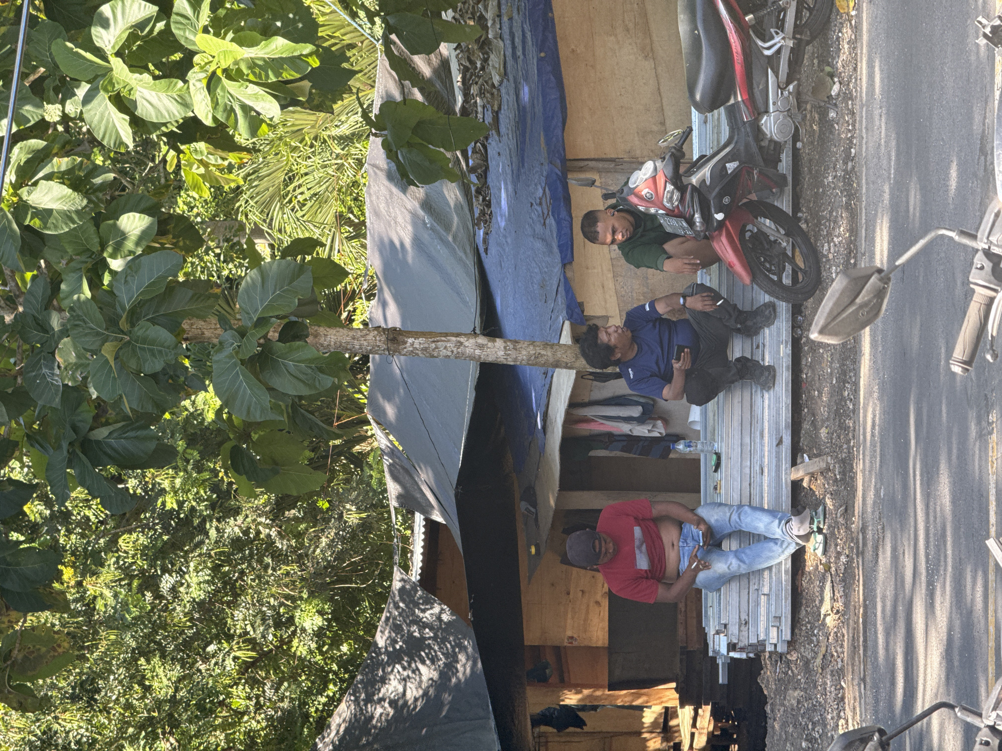 The image, taken in Kuta Selatan, Indonesia, shows three men alongside a road. One man, wearing a red shirt, blue jeans, and a dark cap, is lying on a stack of metal construction materials. Another man, dressed in a blue shirt and black pants, is sitting beside him, looking down. A third man, wearing a green shirt, is sitting near a red and black motorcycle. They appear to be taking a break. A building or structure covered with blue and black tarps is behind them. The roadside environment is visible, including a motorcycle, a paved road, and dense green foliage.