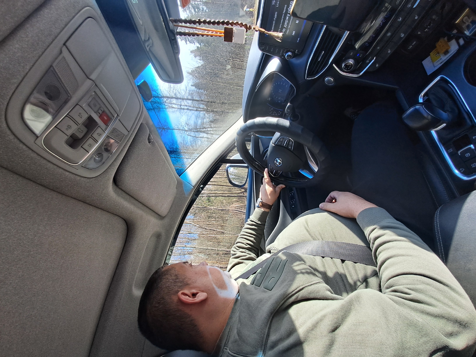 A man is seen reclined in the driver's seat of a Hyundai car. He is wearing a green sweatshirt and a seatbelt. His left hand rests on the steering wheel. Outside the car, trees are visible. The car's dashboard and interior controls are in focus, including the media screen displaying "Bluetooth Audio". The car is likely stationary. No information indicates the location is in Babeevo, Russia.