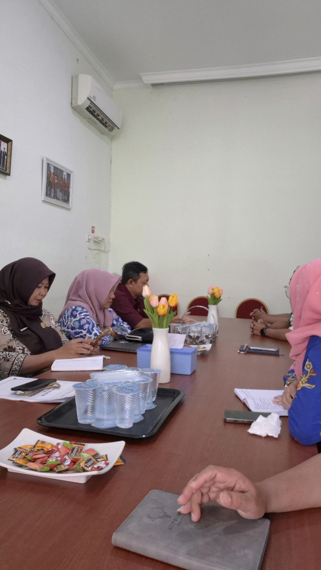 The image depicts an indoor scene, likely a meeting room in Wates, Indonesia. Several individuals are seated around a wooden conference table. On the left, a woman wearing a brown hijab and a patterned top is looking down at her phone. Next to her, a woman in a pink hijab and a blue patterned top is also engaged with her phone, with papers spread out in front of her. A man in a maroon shirt is seated further down the table, facing away from the camera. On the right side of the table, a person wearing a bright pink hijab and a blue embroidered top is visible, as is another individual whose hands are clasped on the table.
On the table, there are several objects: a black tray holding numerous clear plastic cups, a white dish filled with individually wrapped candies, a blue tissue box, and two small vases each containing a bouquet of artificial tulips in shades of yellow and orange. A laptop is also partially visible. In the foreground, a grey notebook with an embossed deer logo rests on the table, with a hand resting on it. In the background, an air conditioning unit is mounted high on a light-colored wall, and two framed pictures are hung on the wall to the left. The lighting suggests an ambient indoor setting.