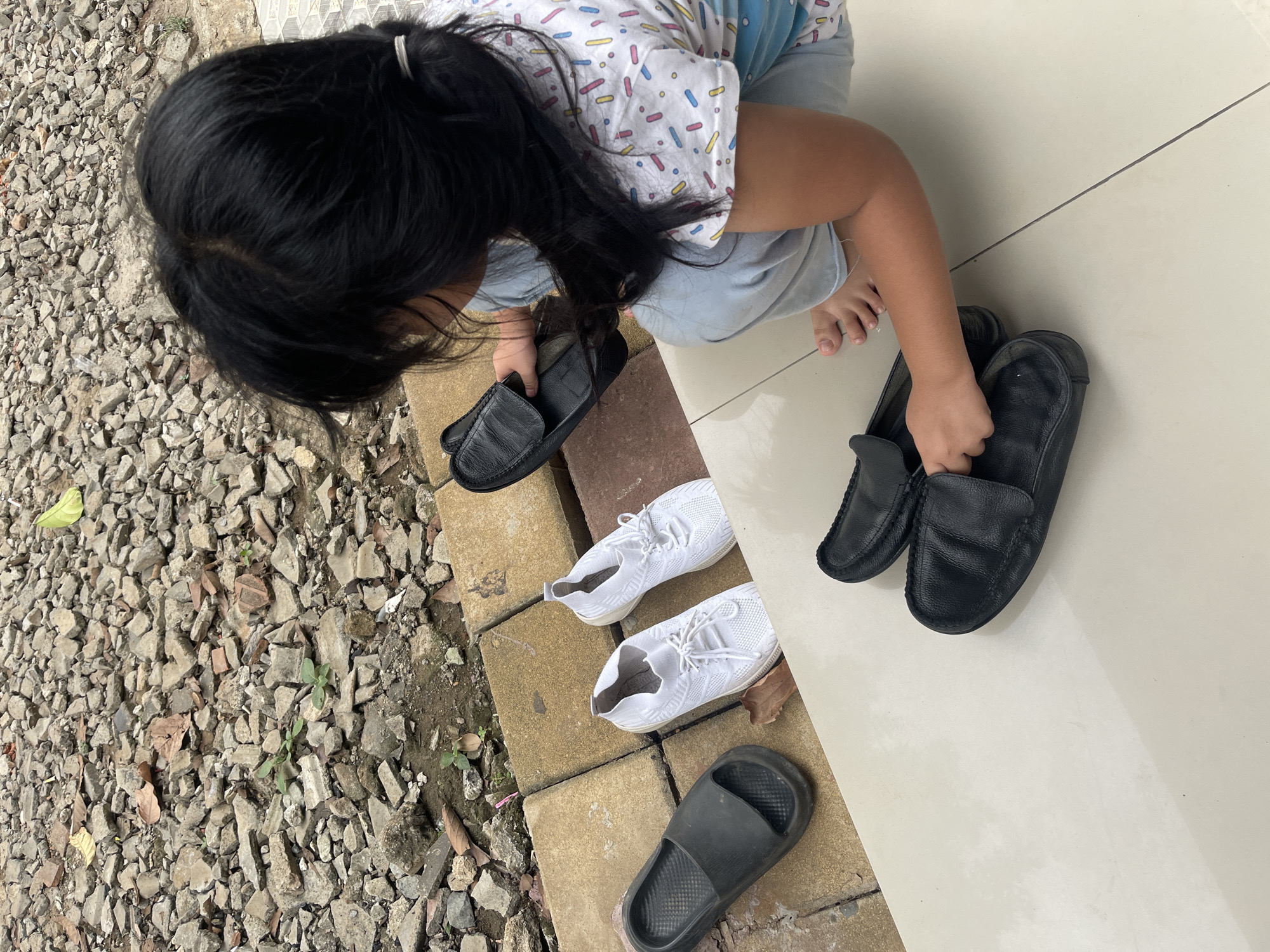 In Gunung Sindur, Indonesia, a young child with black hair is kneeling on a light-colored tiled floor. They are wearing a white t-shirt with colorful abstract patterns and light blue pants. The child is holding a black leather loafer in their right hand, examining it. In front of the child, on the same tiled surface, are two more pairs of black leather loafers, one stacked on top of the other. To the left of these loafers, on a raised platform made of interlocking square tiles, are two white athletic shoes and a single black slide sandal. The background consists of a rough ground surface covered in small rocks and debris, consistent with an outdoor environment in an Indonesian city.