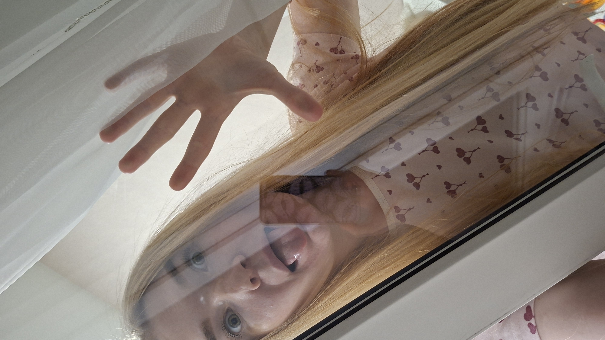 A woman with blonde hair is peering into the camera lens. Her face is close to the camera. Her tongue is partially visible. Her right hand is slightly visible. She is wearing a pink shirt with a small, repeating heart and cherry pattern. A white curtain is in the background. The location is Chaikovsky, Russia.