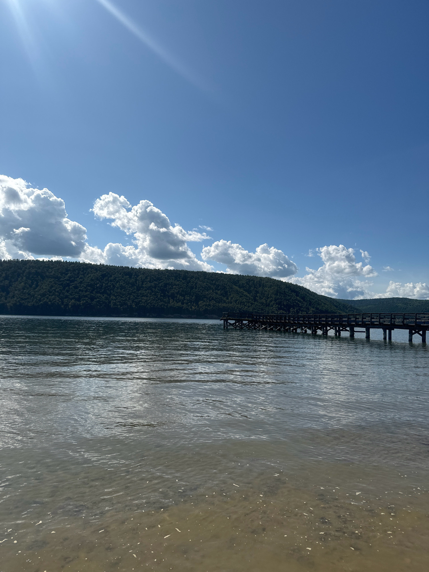 The image depicts a serene natural landscape featuring a body of water, a wooden pier, a tree-covered hillside, and a clear blue sky with scattered white clouds. The foreground shows the shallow, sandy bottom of the water, with ripples on the surface. A dark wooden pier extends from the right side of the image, supported by pilings, and stretches towards the left. Beyond the water, a densely forested hillside rises, its dark green foliage contrasting with the bright sky. The sky is a vibrant blue with fluffy white clouds, and the sun's rays are visible in the upper left corner, creating a bright, luminous effect. The scene appears to be a tranquil outdoor setting, likely a lake or reservoir. The specific location in the city of Тальцы (иркутский), Russia, is not discernible from the image itself, as there are no identifying landmarks or signs.