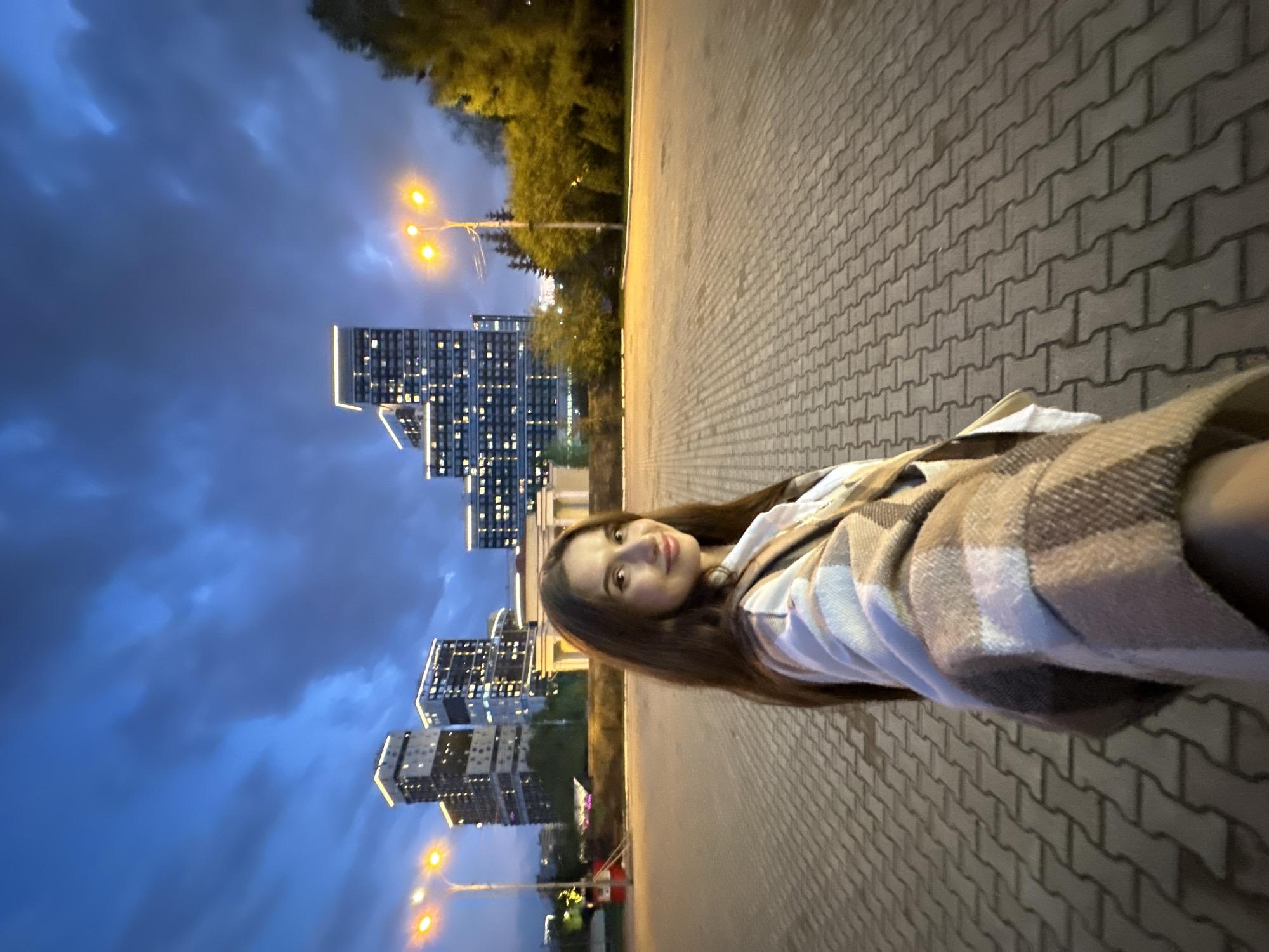 The image shows a young woman standing on a brick-paved sidewalk at twilight. She is wearing a plaid scarf or shawl. Behind her are modern high-rise buildings with illuminated windows, and streetlights are visible. Based on the architectural style of the buildings, the location is likely in Yekaterinburg, Russia. The sky is cloudy and a deep blue color. Trees and greenery can be seen in the background.