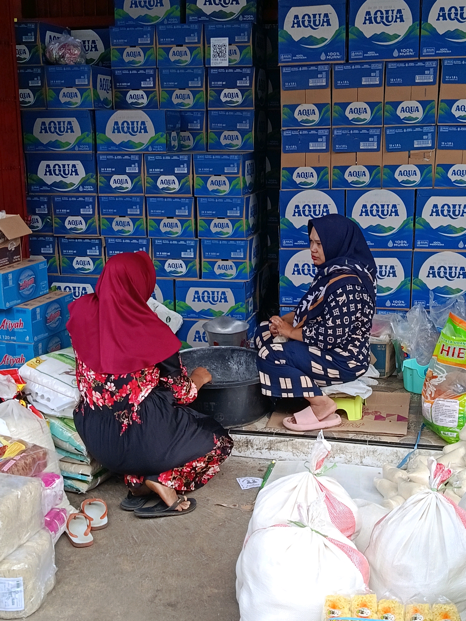 The image depicts an outdoor scene in Sidorenggo, Indonesia, likely a marketplace or storage area, with a prominent display of bottled water crates. Two women are present. The woman on the left, facing away from the camera, wears a red hijab and a floral-patterned dress. She is kneeling beside stacks of rice sacks and a large black tub. To her right, a woman wearing a dark blue hijab and a patterned navy blue outfit is also kneeling. She is facing left and appears to be interacting with the woman on the left. The background is dominated by numerous stacked blue and white crates of "AQUA" bottled water. In the foreground, various bags of goods, including rice and packaged food items, are visible. The ground is paved.
