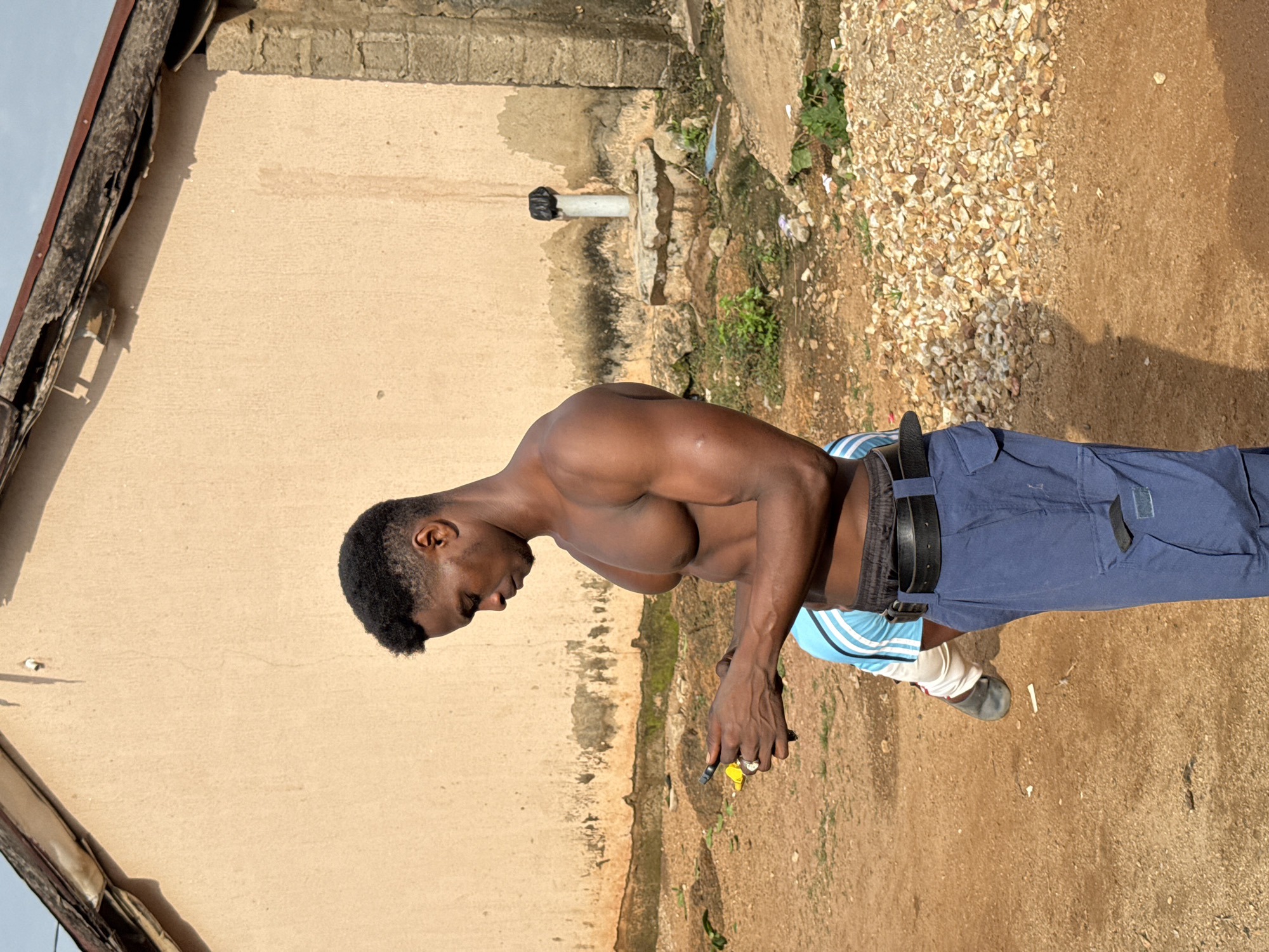 The image shows a shirtless Black man with dark skin and short, black hair standing outdoors in profile, looking down at his hands, which are holding a mobile phone. He is wearing blue cargo pants and a black belt. A portion of another person, wearing a blue and white striped shirt and white pants, is visible behind him. The background consists of a tan-colored building with a damaged roofline, a pile of gravel, and a patch of dry, reddish-brown earth. There are also some green plants and a concrete structure with a white pipe and black cap. The lighting suggests it is daytime, with shadows cast by the subjects and building. The scene appears to be in a semi-urban or rural area, consistent with Akwanga, Nigeria.