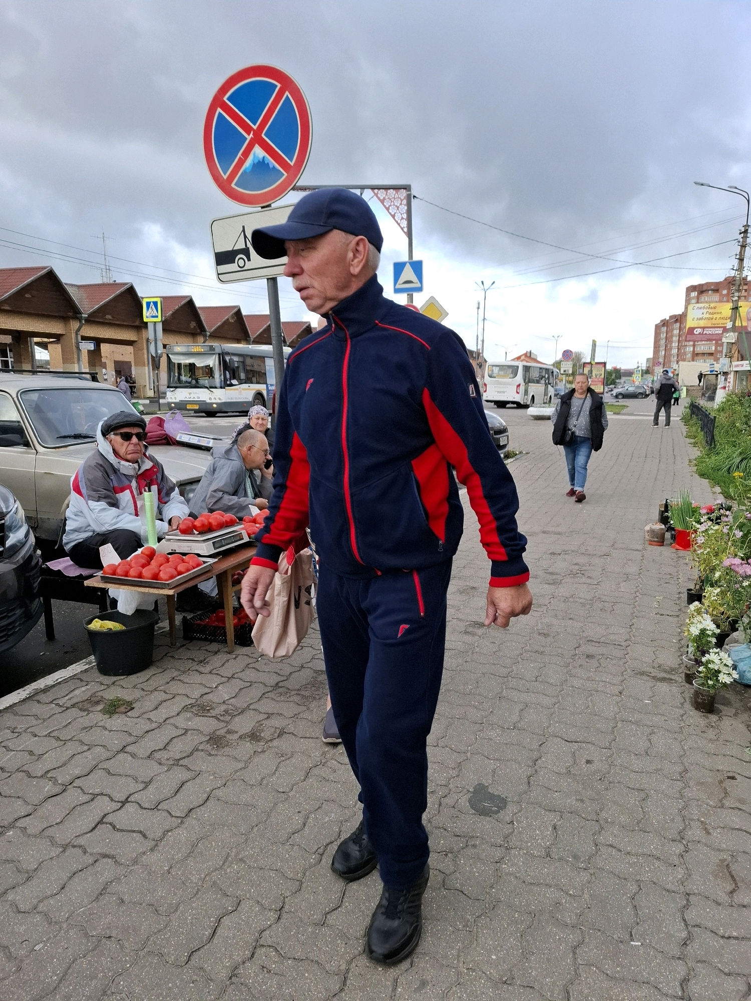 The image captures an outdoor market scene under an overcast sky. In the foreground, a man wearing a blue and red tracksuit and a blue baseball cap walks towards the right. Behind him, two men are seated at a table displaying a large pile of tomatoes. One of them, wearing a grey and white jacket and sunglasses, is looking towards the left. The other man, in a grey jacket, is on the phone. A black bucket with a yellow object is on the ground next to the table. A car is parked to the left of the scene. Further in the background, several buildings with red-tiled roofs are visible. A bus is stopped at a bus stop, and people are walking on the sidewalk. Traffic signs, including a "no parking" sign, are present. The ground is paved with interlocking grey bricks. Flower pots with various plants are arranged along the right side of the sidewalk. The overall impression is of a functional, everyday street market.