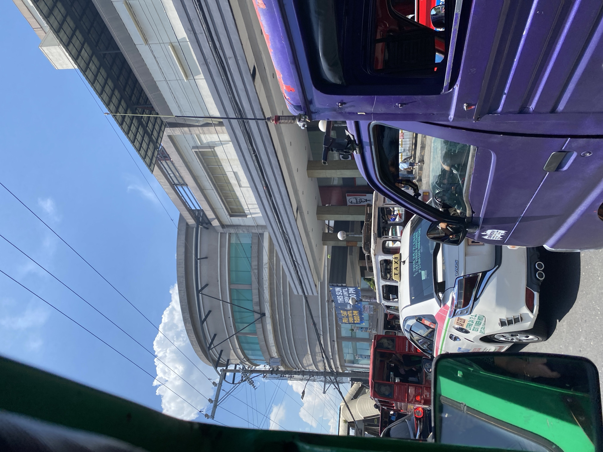 The image captures a vibrant street scene in Davao City, Philippines, under a bright blue sky with scattered white clouds. The photograph is taken from inside a green vehicle, possibly a tricycle or jeepney, with its front frame partially visible in the foreground.
The street appears to be busy with traffic. In the mid-ground, a prominent purple jeepney occupies the right side, showing its robust design and a long antenna extending upwards. Following it is a white Toyota Vios taxi, clearly marked with "TAXI" on its roof. Details on the taxi include "VIOS" on the trunk, "LTFRB 24/7:1342" and "TEXT/VIBER HOTLINE 0917-550-1342 / 0998-550-1342" printed on its side. A red vehicle, possibly another jeepney, is visible behind the taxi, with text like "MARIANA" and a small sticker displaying "740" with an indistinct symbol. Reflections of people can be seen in the vehicle windows, though no distinct faces are visible.
On the left side of the street, a modern, multi-story building with a distinctive curved facade and green-tinted windows stands out. Further down the street, other commercial buildings line the road, one faintly showing a KFC logo. Numerous black power lines crisscross the sky, connecting to utility poles that also support a blue banner. The banner displays business hours: "FRI-SAT OPEN UNTILL 10:00 PM" and "THIS SUN OPEN UNTIL 1:00 PM". The scene depicts a typical bustling daytime environment in a Philippine urban center.