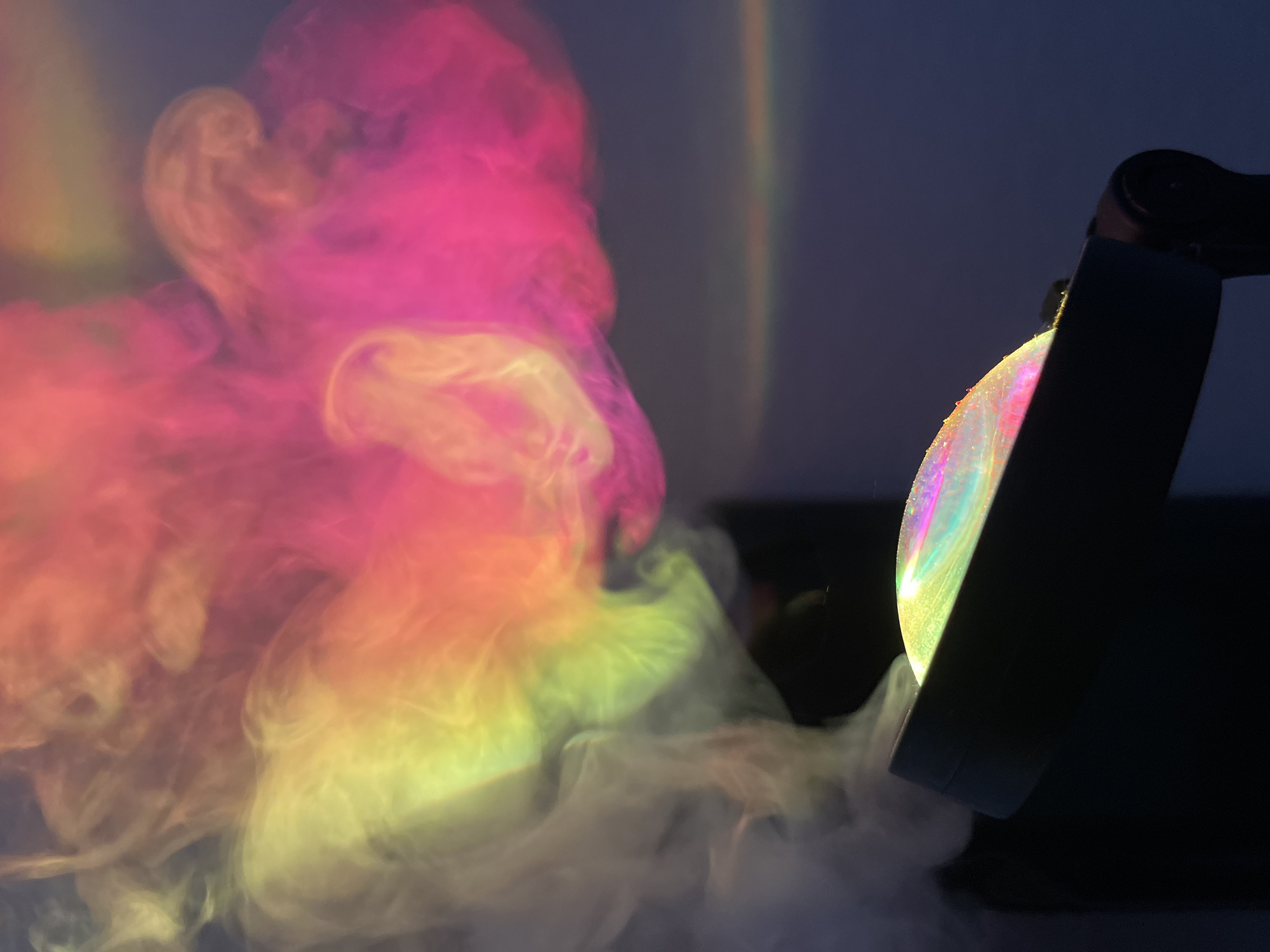 The image shows a light projector casting colored light onto smoke. The projector is dark in color and has a round lens with rainbow-like colors. The smoke is illuminated by the light, creating a colorful, swirling effect with shades of pink, orange, yellow, and white. The background is a solid dark color, possibly a wall. The location is Unknown, Unknown.