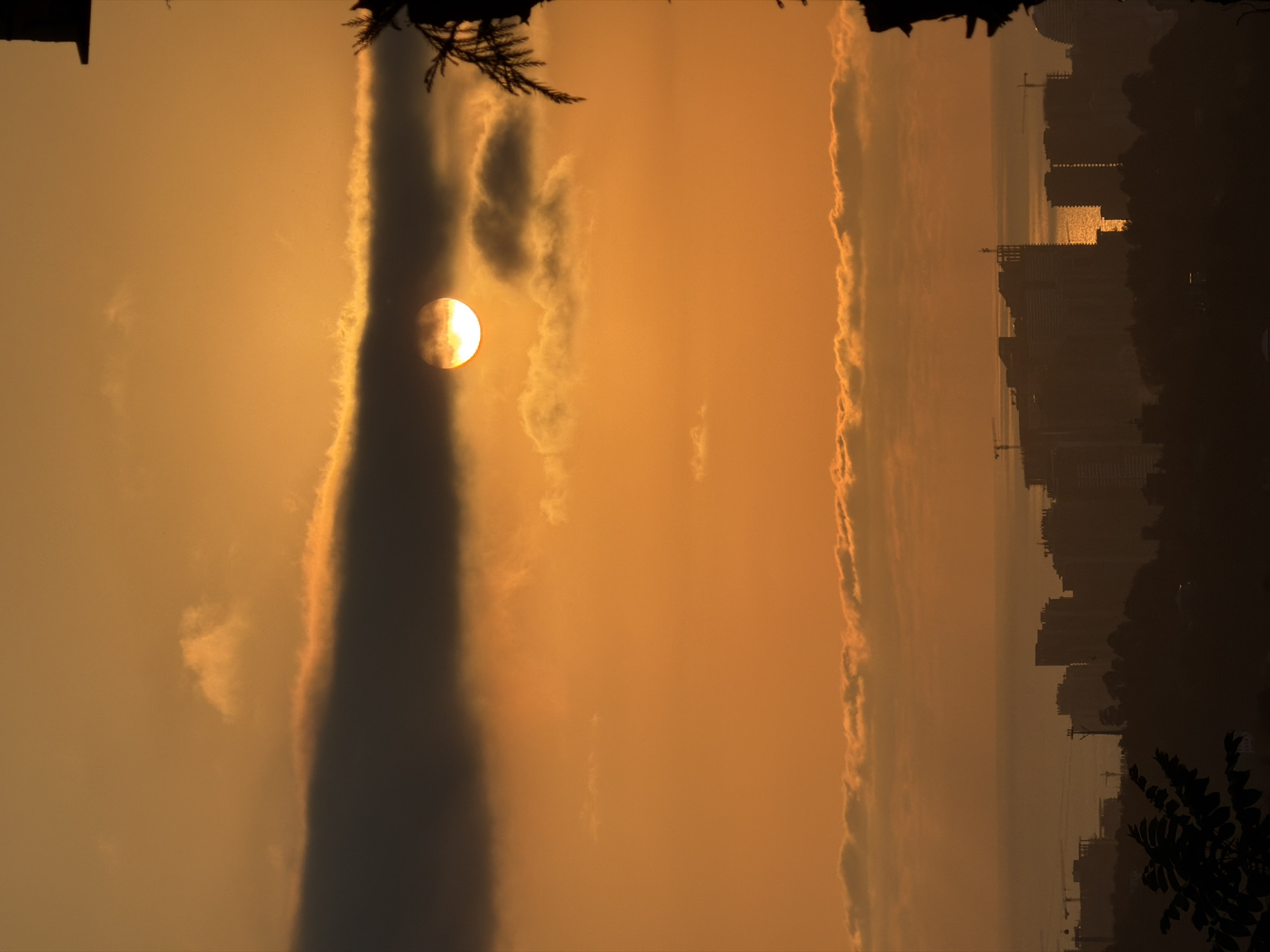 The image depicts a sunset over Akhalsheni, Georgia. The sun is partially obscured by dark clouds in the upper portion of the frame, casting a warm orange glow across the sky. Below the clouds, a line of lighter, fluffy clouds stretches horizontally. In the distance, a silhouette of buildings and construction cranes is visible against the horizon, suggesting an urban or developing area. The sea or a large body of water can be seen behind the city skyline. The foreground features dark, silhouetted foliage, possibly trees or branches, framing the scene on the right and left sides. The overall impression is one of a developing cityscape at dusk.