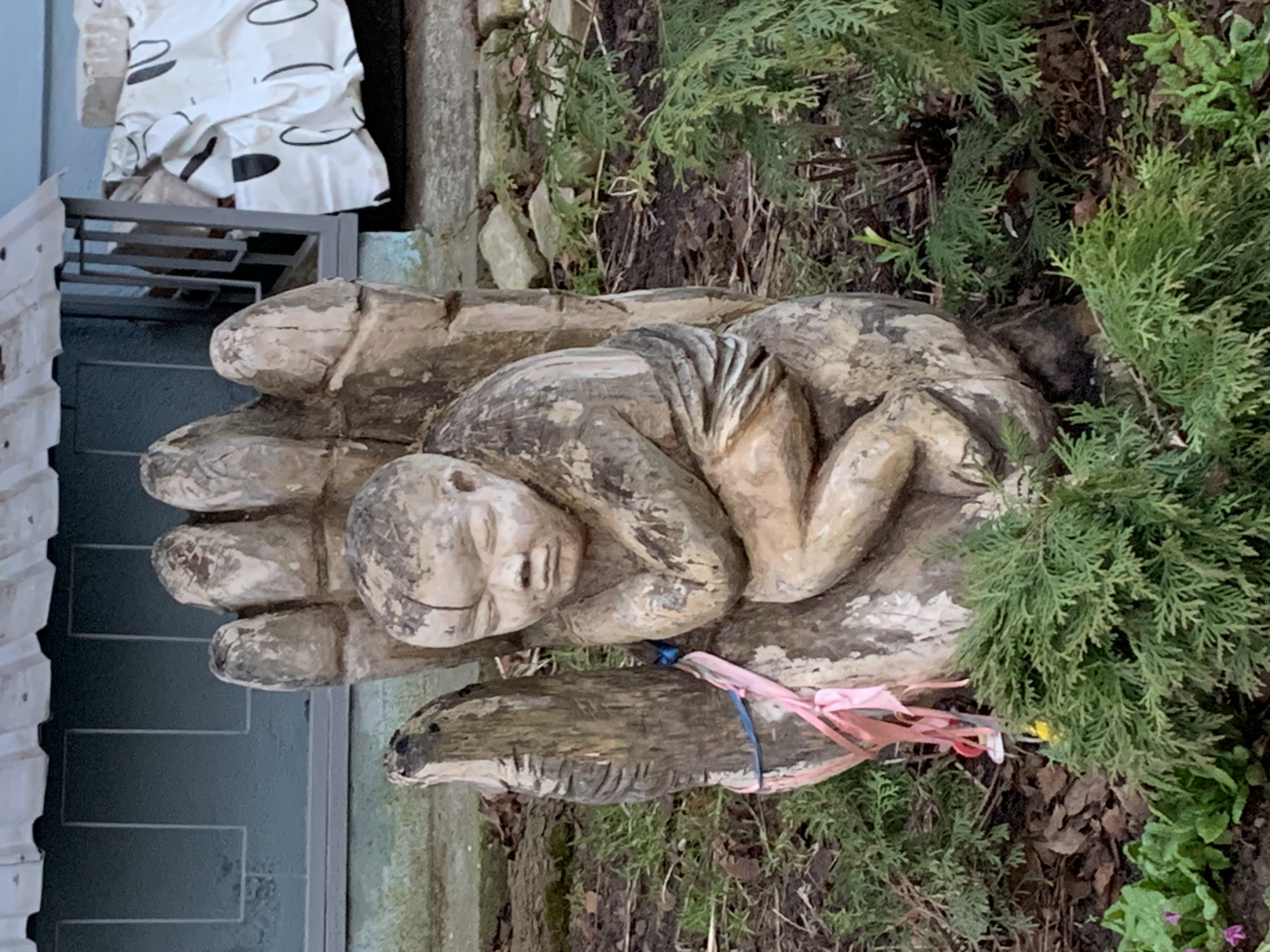 The image shows a wooden sculpture depicting a person lying in the palm of a hand. The sculpture is weathered and appears to be outdoors, situated in a garden setting with greenery, plants, and ground cover surrounding it. A blue string and pink ribbons are tied around the sculpture. In the background, parts of a building are visible. Based on the available information, it is a sculpture in Chaikovsky, Russia.