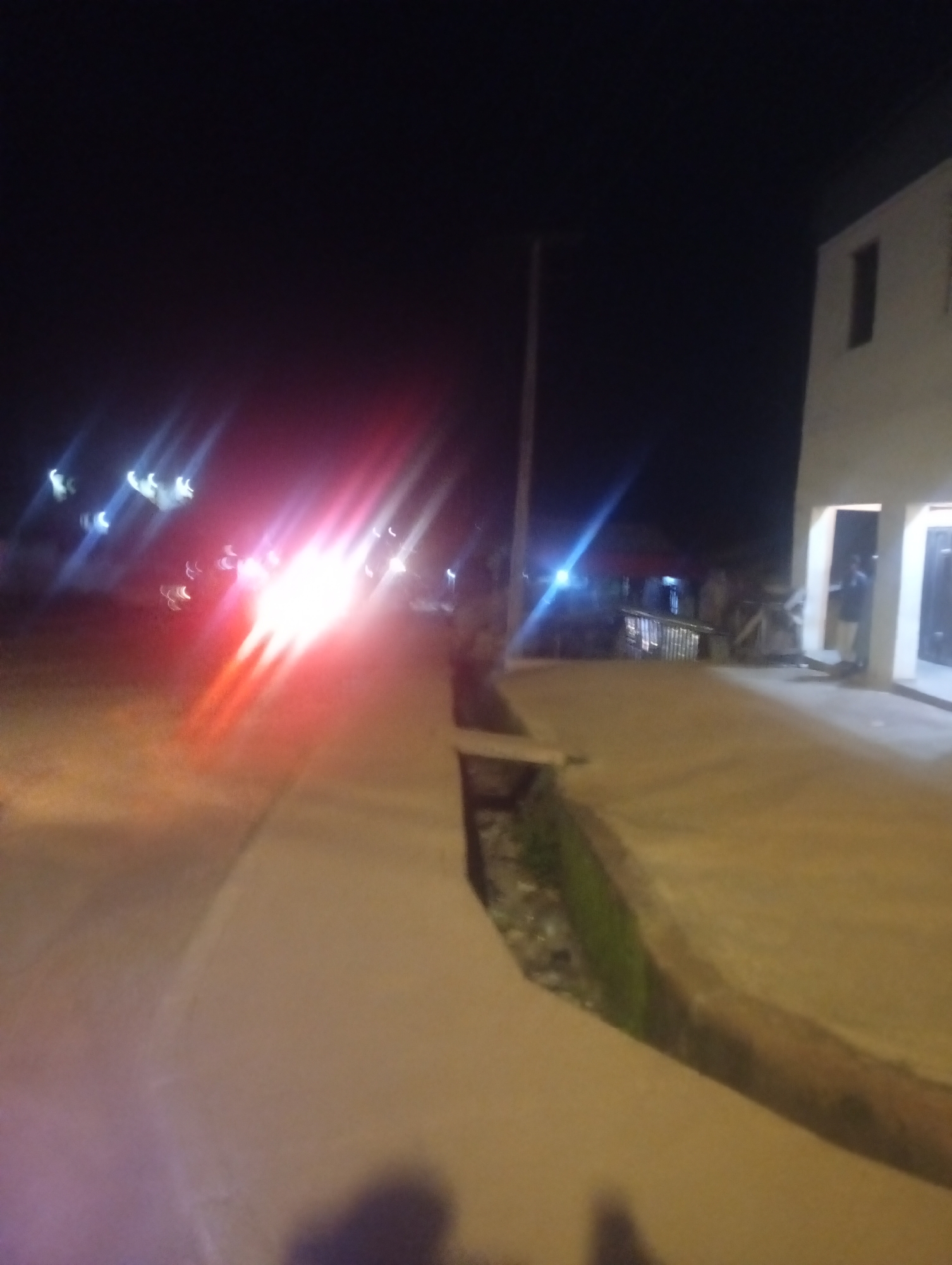 The image depicts a nighttime scene on a street in Bukuru, Nigeria. The primary visual element is the bright, blurred lights of a vehicle or vehicles approaching from the left, casting red and white streaks across the asphalt. To the right, a portion of a light-colored building is visible, with an open doorway. The ground appears to be a paved walkway or road with a drainage ditch running alongside it, containing some greenery. The overall impression is one of low light conditions with significant light trails due to motion or camera settings. There are indistinct figures visible in the doorway of the building.