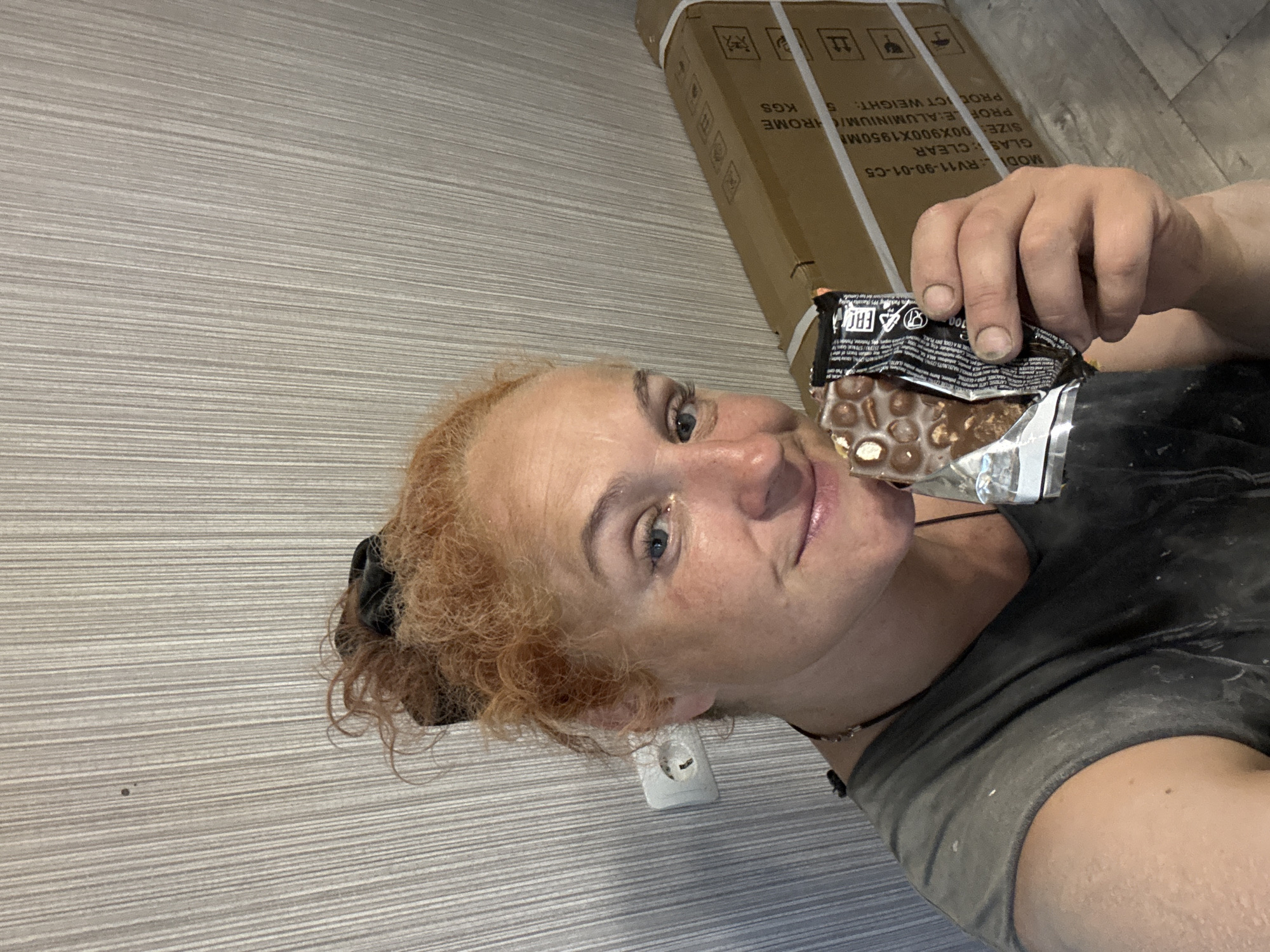 In Chaikovsky, Russia, a woman with red hair tied up with a black scrunchie holds a chocolate bar. She is wearing a dark gray t-shirt, which appears to be dusty or stained, suggesting a work environment. Her left hand, with dirt under the fingernails, is holding the unwrapped chocolate bar. In the background, a large cardboard box is visible, strapped with white tape, and a wall with a striped texture. A white electrical outlet is mounted on the wall to the left of the woman. The floor is a light gray material. The overall scene appears to be indoors, possibly in a construction or renovation setting, given the dust and the box.