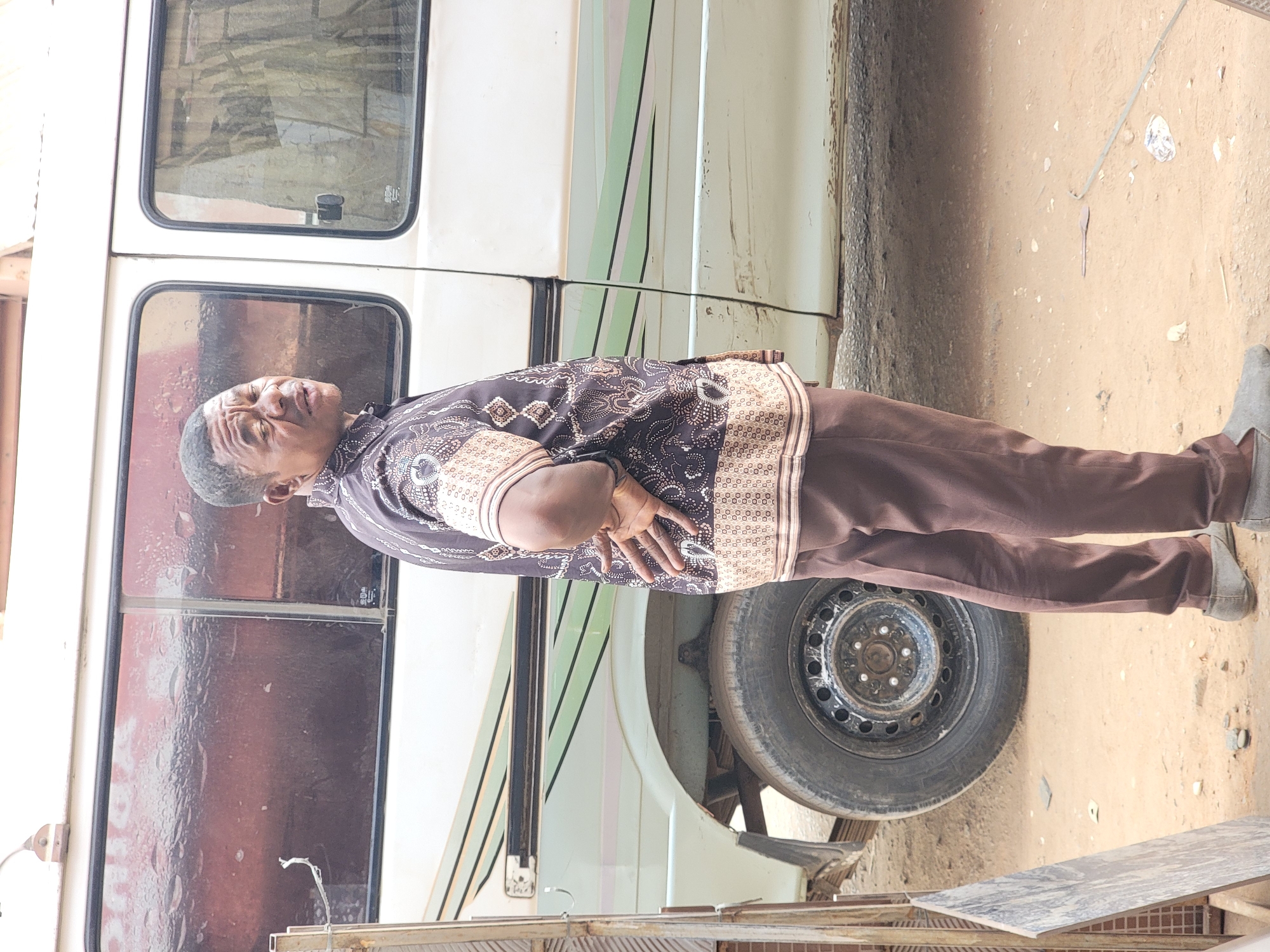An adult male stands with arms crossed in front of a light-colored van. He wears a patterned brown and beige shirt and brown pants. The location appears to be outdoors, potentially in Maiburiji, Nigeria. The van is positioned near a wall and a dirt ground.