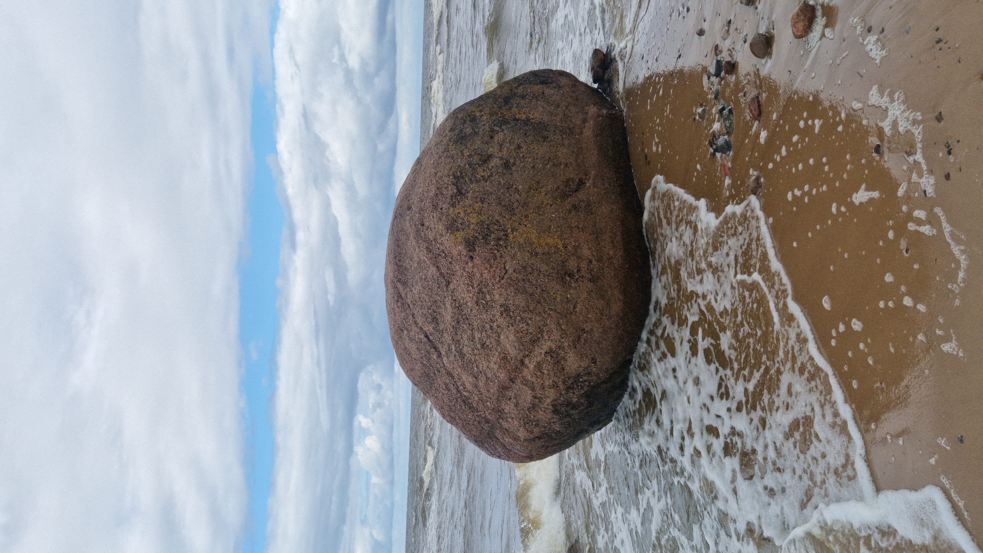 A large, rounded, reddish-brown boulder rests on a sandy beach, partially submerged by the gentle lapping of waves. The water is a muted greyish-blue, with small whitecaps visible in the distance. The sky above is overcast with a mix of white and grey clouds, with patches of pale blue sky peeking through. The sand is wet and reflects the light, with a scattering of smaller pebbles and shells. In the background, the sea stretches towards the horizon. The scene appears to be a natural landscape, likely a coastal area. The image was taken in Vārzas, Latvia.