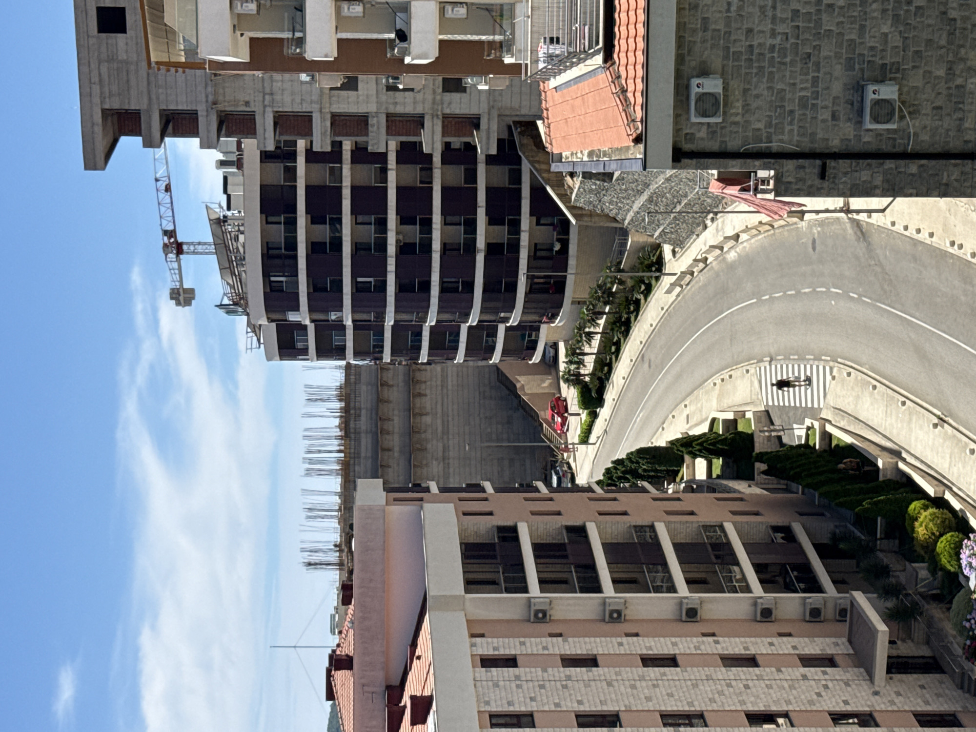 This is an aerial, eye-level, medium shot of a construction site with several apartment buildings in Boreti, Montenegro. The sky is blue with some wispy clouds. In the foreground, a light pink apartment building with a tiled roof and balconies is on the left. To the right of this building is a curved road with a single person walking on a crosswalk. Further back, a dark-colored, curved apartment building is under construction, with a tall construction crane visible above it. Behind this building is a massive, concrete structure with rebar protruding from the top, indicating ongoing construction. Another apartment building, partially constructed with concrete and brick, is on the far right. The scene depicts a developing urban environment with a blend of completed and under-construction residential properties.