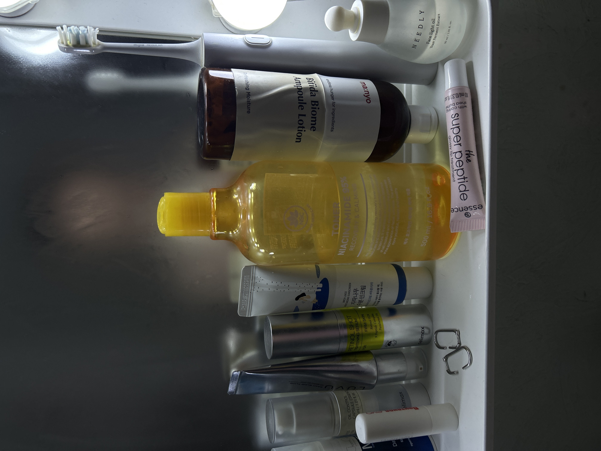 A high-angle, medium shot captures a collection of skincare products arranged on a white shelf against a metallic, reflective background. In the foreground, from left to right, are a white tube with a silver cap labeled "OIL CONTROL CAPSULE ESSENCE," followed by a silver tube with a blue and white label. Next is a slender silver tube labeled "RETINOL SHOT FIRM SKIN," with text indicating "Retinol 0.2%." To its right is a white tube with a blue and white illustrated design and Korean text, followed by a large yellow bottle of toner. A brown bottle labeled "Bifida Biome Ampoule Lotion" stands behind the yellow toner. To the right of the brown bottle is a white electric toothbrush. Further to the right is a clear bottle labeled "NEEDLY Face light oil Yeast Ferment Extract." A small pink tube labeled "essence the super peptide" lies horizontally in front of the other items. A pair of silver hoop earrings are placed near the center of the shelf. The overall scene suggests a curated personal care collection, likely in a bathroom or vanity setting.