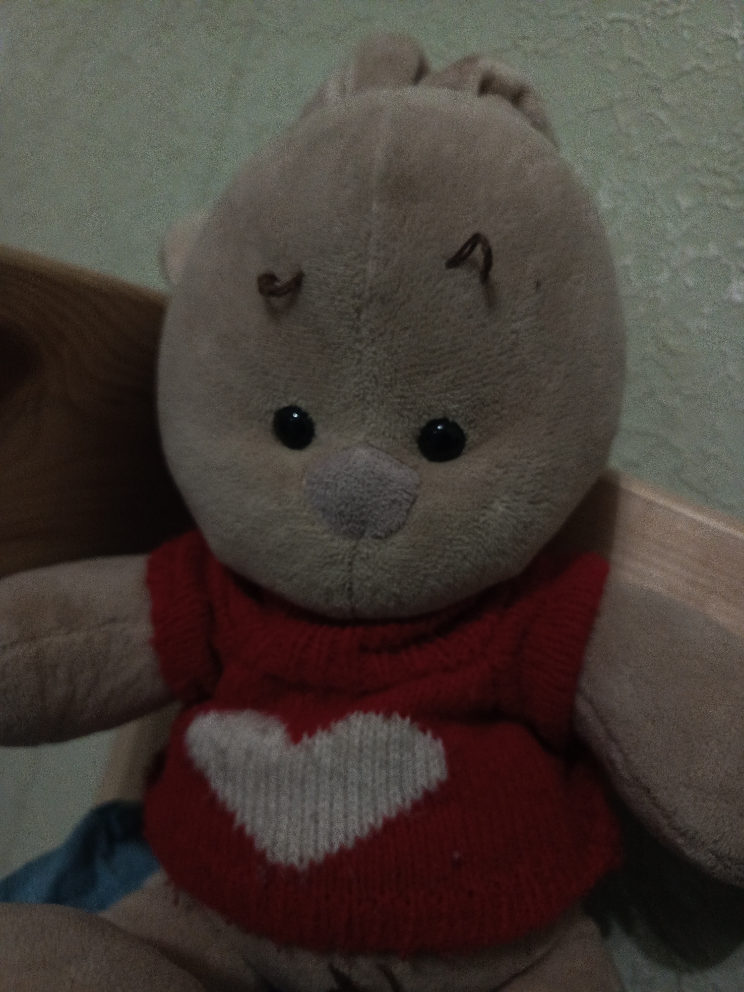 A close-up shot of a beige stuffed animal wearing a red knitted sweater with a white heart on the front. The stuffed animal has black eyes, a gray nose, and two brown yarn eyebrows. The background is a pale green wall. The location is unknown.