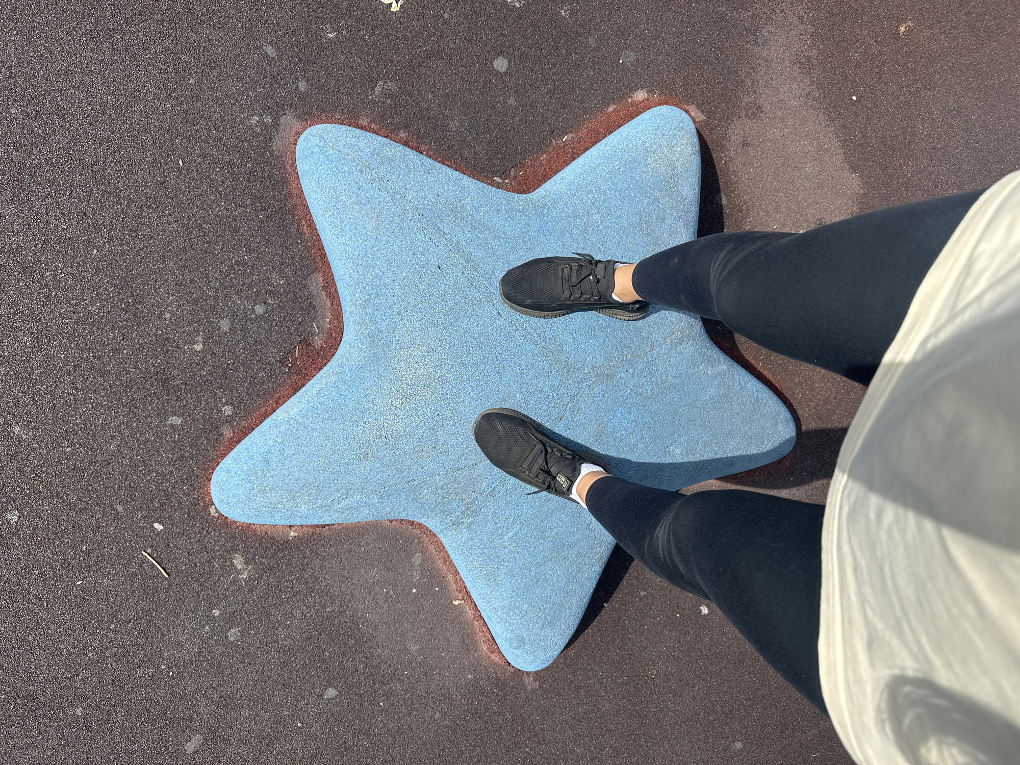 A high-angle shot captures a person standing on a blue star-shaped playground feature. The person is wearing black leggings and black sneakers with white socks. They are positioned in the center of the star, with their legs and torso visible from the knees up. The star is situated on a dark, reddish-brown rubberized playground surface. The lighting suggests a sunny day, casting a shadow to the right of the star. The image does not contain any elements that would allow for identification of its location within Petropavlovsk-Kamchatsky, Russia.