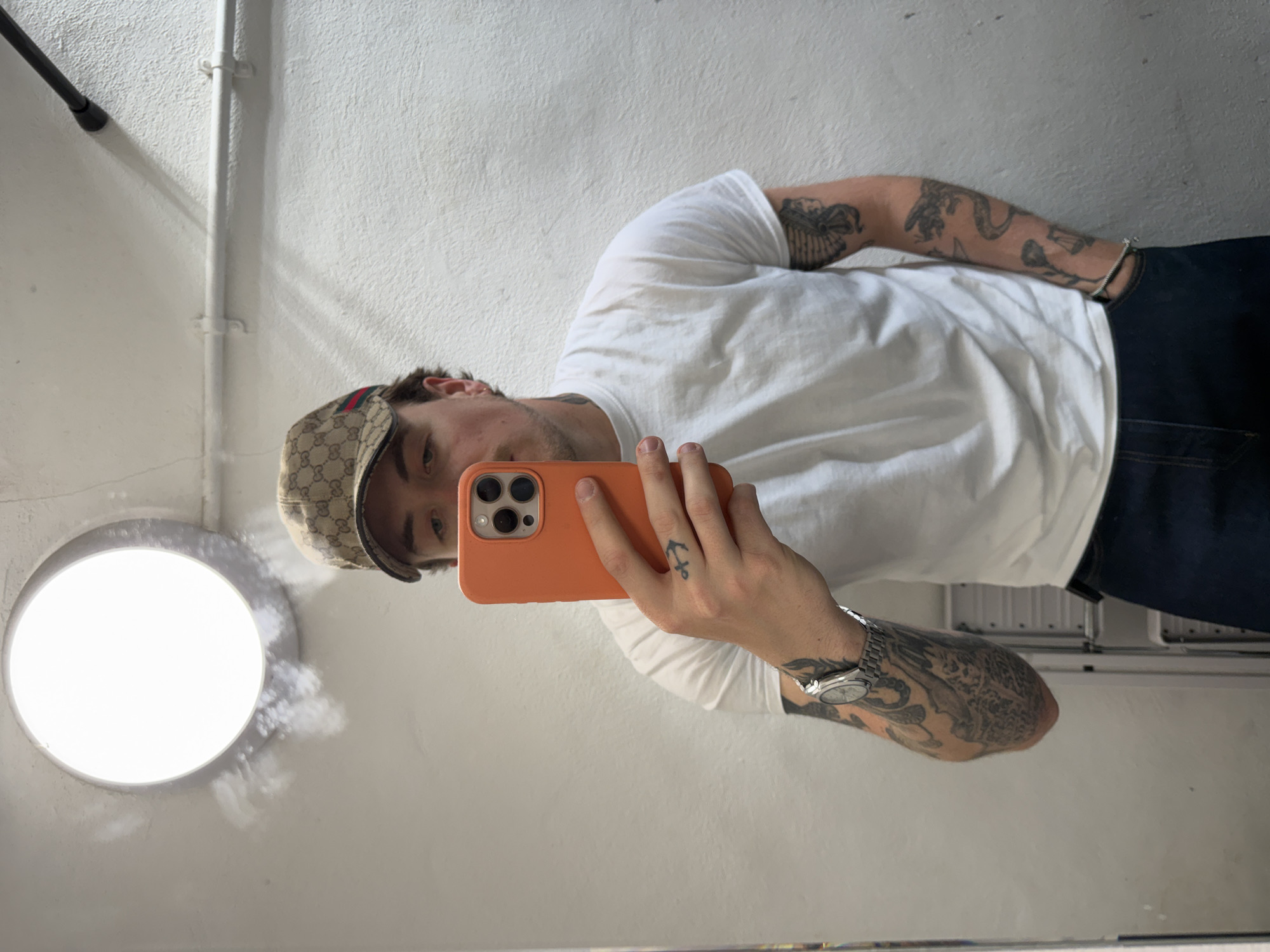 A man takes a selfie in a mirror. He wears a patterned cap, a white t-shirt, and dark pants. His arms are tattooed, and he wears a watch and a bracelet. The phone he uses for the selfie has an orange case. The background consists of a white wall, a light fixture, and a pipe. There is no information to deduce that this image was taken in London.