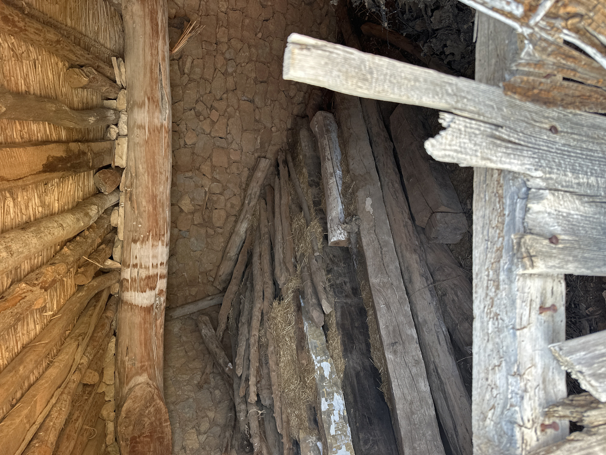 The image shows the interior of a rustic structure, likely an old barn or shed, located in Alpuente, Spain. The scene is dominated by raw, weathered wood and stone construction.
The ceiling is made of rough-hewn wooden beams and smaller branches, creating a rustic and uneven surface. A large, thick beam runs horizontally across the middle of the image, supporting the ceiling structure and resting against a stone wall. This beam shows signs of wear and discoloration.
The back wall is constructed of rough-cut stones, laid in an irregular pattern. Piles of chopped logs and branches are stacked against this wall and fill much of the floor space. Some of the wood appears to be partially covered by dry straw or hay. The logs vary in size and condition, with some appearing freshly cut and others weathered and aged.
In the foreground, more wooden beams and planks are visible, some of which appear to be part of a damaged or decaying structure, possibly a fence or partition. These foreground elements are also made of aged, greyed wood, some showing signs of splitting and damage. Small, rusty nails are visible in some of these wooden pieces.
The overall impression is one of disuse and decay, a scene of agricultural storage or a place of shelter that has fallen into disrepair. There are no people or animals present in the image. The lighting suggests it is daytime, with natural light filtering into the interior, creating shadows and highlighting the textures of the materials.