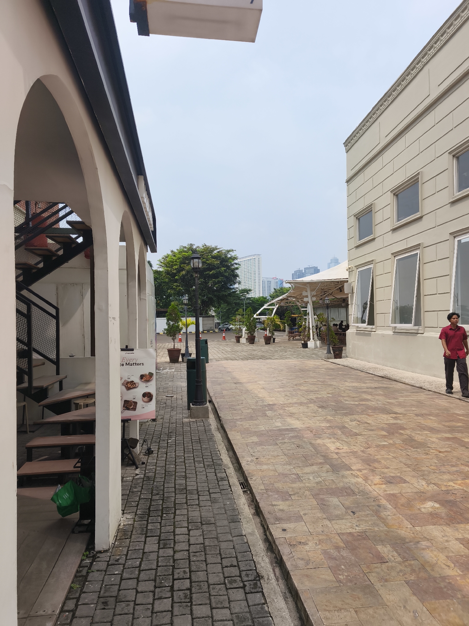 The image depicts an outdoor scene with a paved walkway lined by buildings and greenery. The architecture suggests a commercial or mixed-use development. On the left, there's a building with arched entrances and an outdoor staircase. A sign visible on the left reads "Every te Matters." On the right, a light-colored, modern building with large windows runs along the walkway. A man in a red shirt and dark pants is walking on the right side of the walkway. Further down the walkway, there are potted plants, outdoor seating areas, and awnings. In the background, tall buildings indicate an urban setting, consistent with South Jakarta, Indonesia. The sky is overcast, suggesting a daytime scene with diffused light.