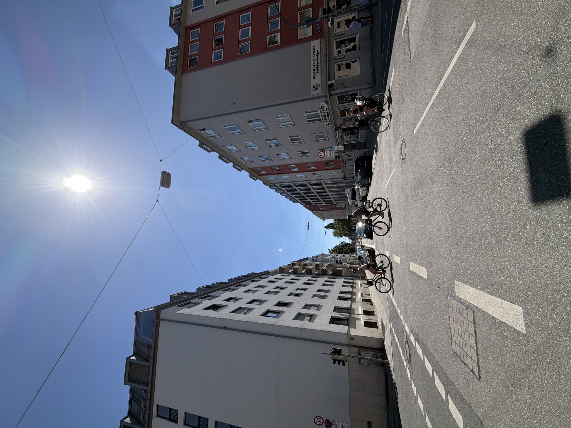 The image shows a street scene in Munich, Germany, on a bright, sunny day with the sun directly overhead. Two apartment buildings line opposite sides of the street. The building on the left is white with multiple floors and many windows. The building on the right is gray with red accents and also has multiple floors and numerous windows.
In the foreground, there are two cyclists on the road. The cyclist on the left is wearing a light-colored top and riding a bicycle. The cyclist on the right is wearing a darker top and is also on a bicycle. Both cyclists are moving away from the viewer.
The street is paved and marked with white lines indicating lanes and crosswalks. There are traffic lights and street signs visible, including a speed limit sign (30). Several cars are parked or driving on the street in the background. There is also a manhole cover in the road. The sky is clear and blue. The overall impression is a typical urban street scene in Germany during the day.