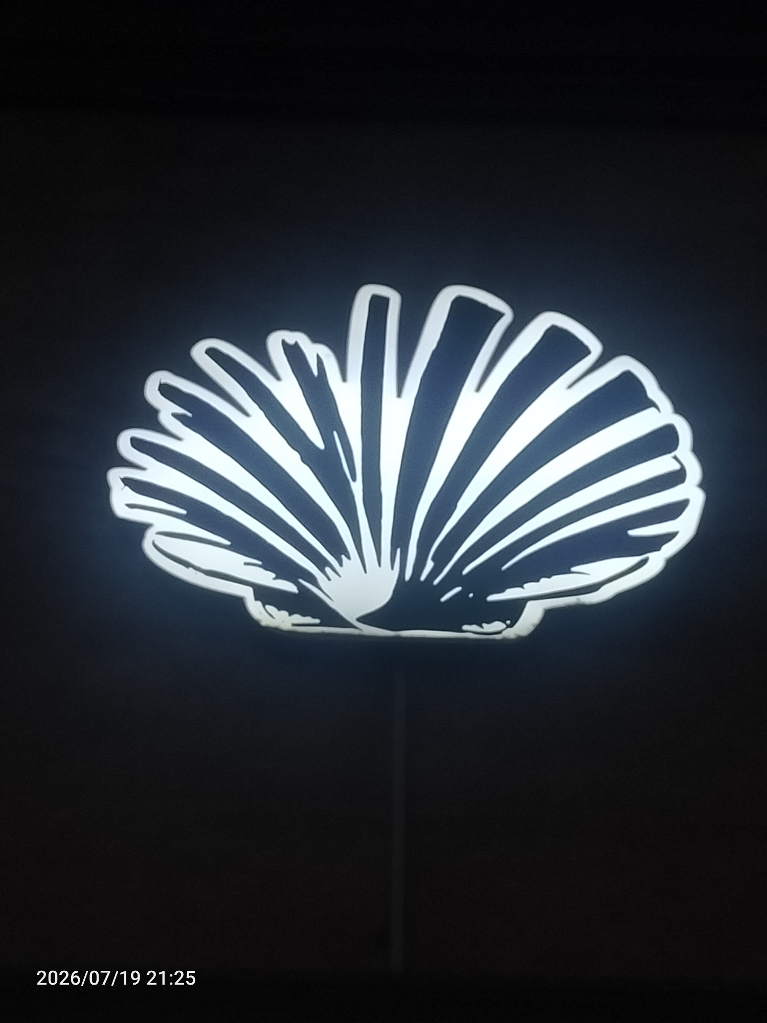 A stylized, illuminated shell-shaped object is centered against a dark background. The shell is white with dark gray or black shading to create a ribbed effect. A thin, vertical pole extends downwards from the bottom center of the shell. The timestamp "2026/07/19 21:25" is visible in the bottom left corner. The scene appears to be indoors or in a very dark outdoor environment.