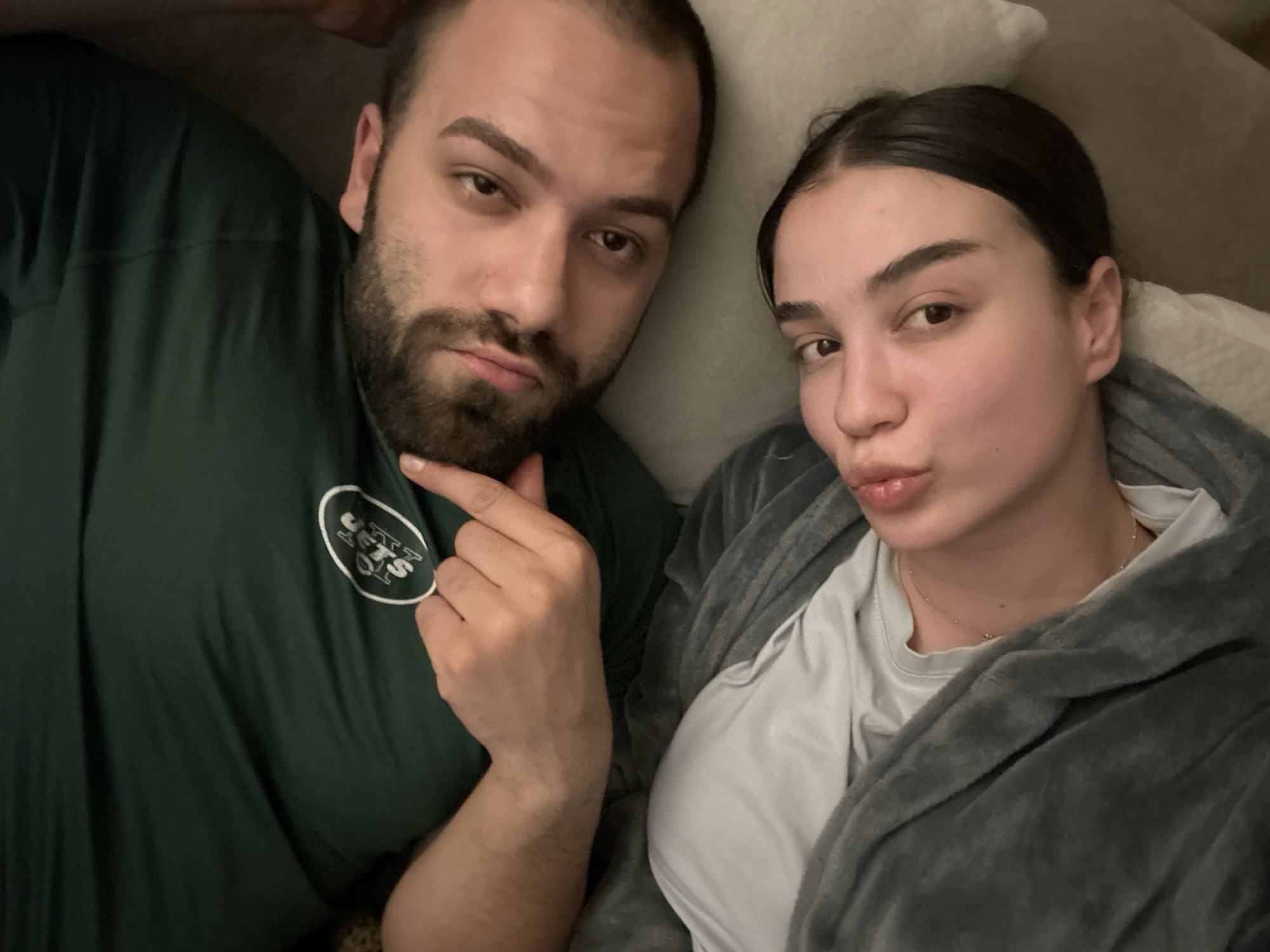 A selfie taken from above features two individuals indoors, seemingly relaxed and potentially in a domestic environment. On the left, a man with a beard, short dark hair, and olive skin wears a dark green shirt with a logo. He is holding his hand up with his finger touching his chin. To the right of him, a woman with dark hair pulled back and light skin is wearing a light-colored shirt with a gray fuzzy robe or blanket over it. Her lips are puckered in a kissing expression. The background consists of pillows. There is nothing to indicate this image was taken in Sofia, Bulgaria.