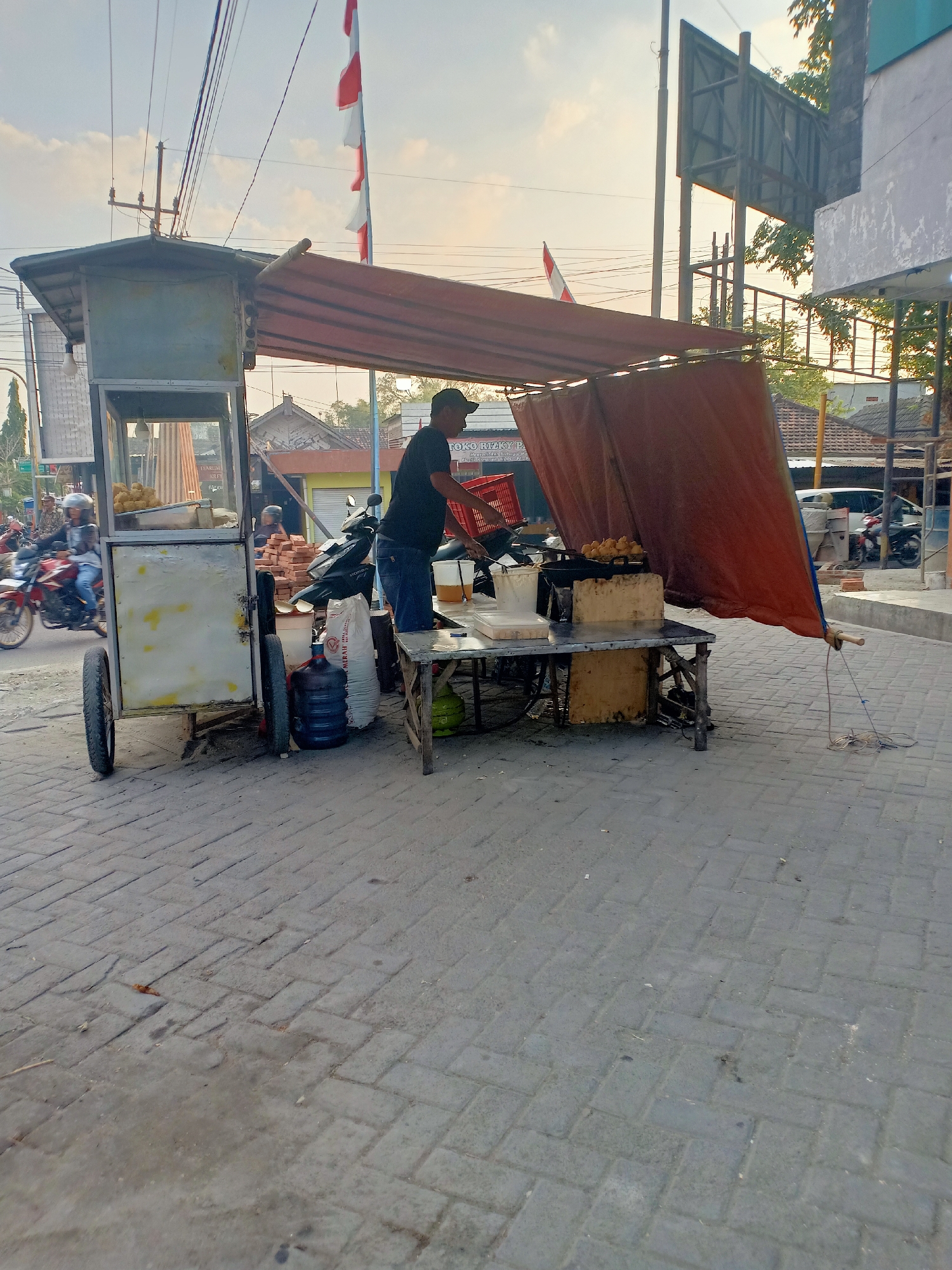 A street vendor is frying food at a cart on a brick-paved street in Dander, Indonesia. The vendor, a man wearing a dark t-shirt and jeans, stands behind a portable cart with a red tarp covering the top. He is actively cooking in a large pan on a metal table. Several large water bottles and sacks are stacked near the cart, and a gas cylinder is visible under the table. In the background, motorcycles and pedestrians are moving along the street, and buildings with tiled roofs are visible. Indonesian flags are flying from poles in the distance. The lighting suggests it is either early morning or late afternoon.