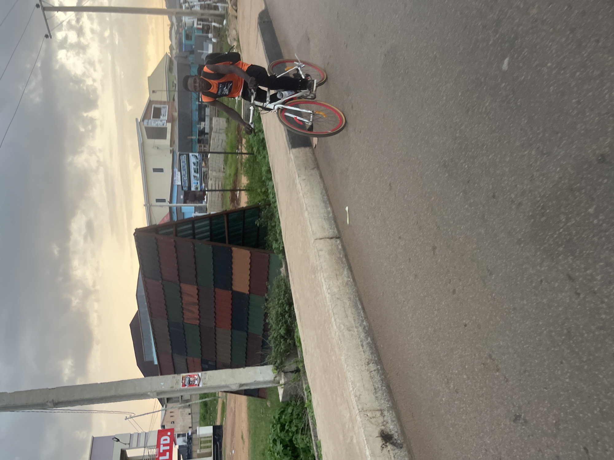 A young man with dark skin and short dark hair is riding a white bicycle with pink rims on a paved road. He is wearing an orange sleeveless t-shirt with a graphic on the front and black pants, with a black backpack on his back. The man is looking towards the camera. The bicycle has a water bottle mounted on the frame. He is positioned on the right side of the road, which appears to be in an urban or suburban area.
In the background, to the left, is a light-colored building with a red sign that says "LTD." A concrete utility pole stands on the left side of the frame, with some posters attached to it. Adjacent to the road is a sidewalk with a concrete curb. Behind the sidewalk, there is a grassy area with some bushes and a large, triangular structure covered in multi-colored roofing tiles. Further back, several buildings are visible, including one with a light-colored facade and a sloping roof, and others that are more industrial in appearance. The sky is cloudy and appears to be either early morning or late afternoon, with a soft, diffused light. The overall setting suggests a location in Gura Top, Nigeria, given the visual context.