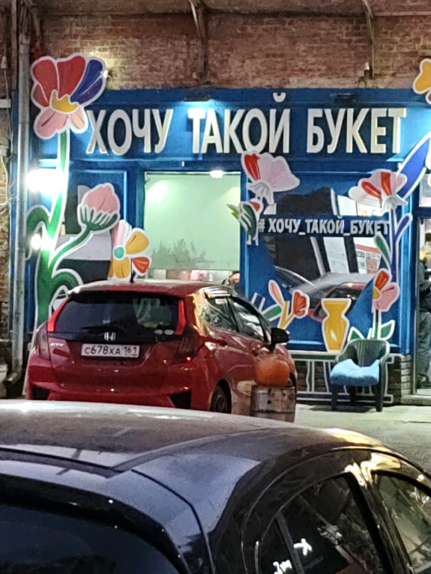 The image depicts a storefront with a large blue sign that reads "ХОЧУ ТАКОЙ БУКЕТ" (I WANT SUCH A BOUQUET) in white, stylized lettering. The sign is adorned with colorful floral illustrations and has a hashtag "#ХОЧУ_ТАКОЙ_БУКЕТ" below it. In front of the storefront, a red Honda Fit car with the license plate "C678XA 161" is parked. To the right of the car, a blue armchair sits on a patio. The background shows a brick building and other vehicles. The scene appears to be a street in Rostov-on-Don, Russia, possibly a flower shop.