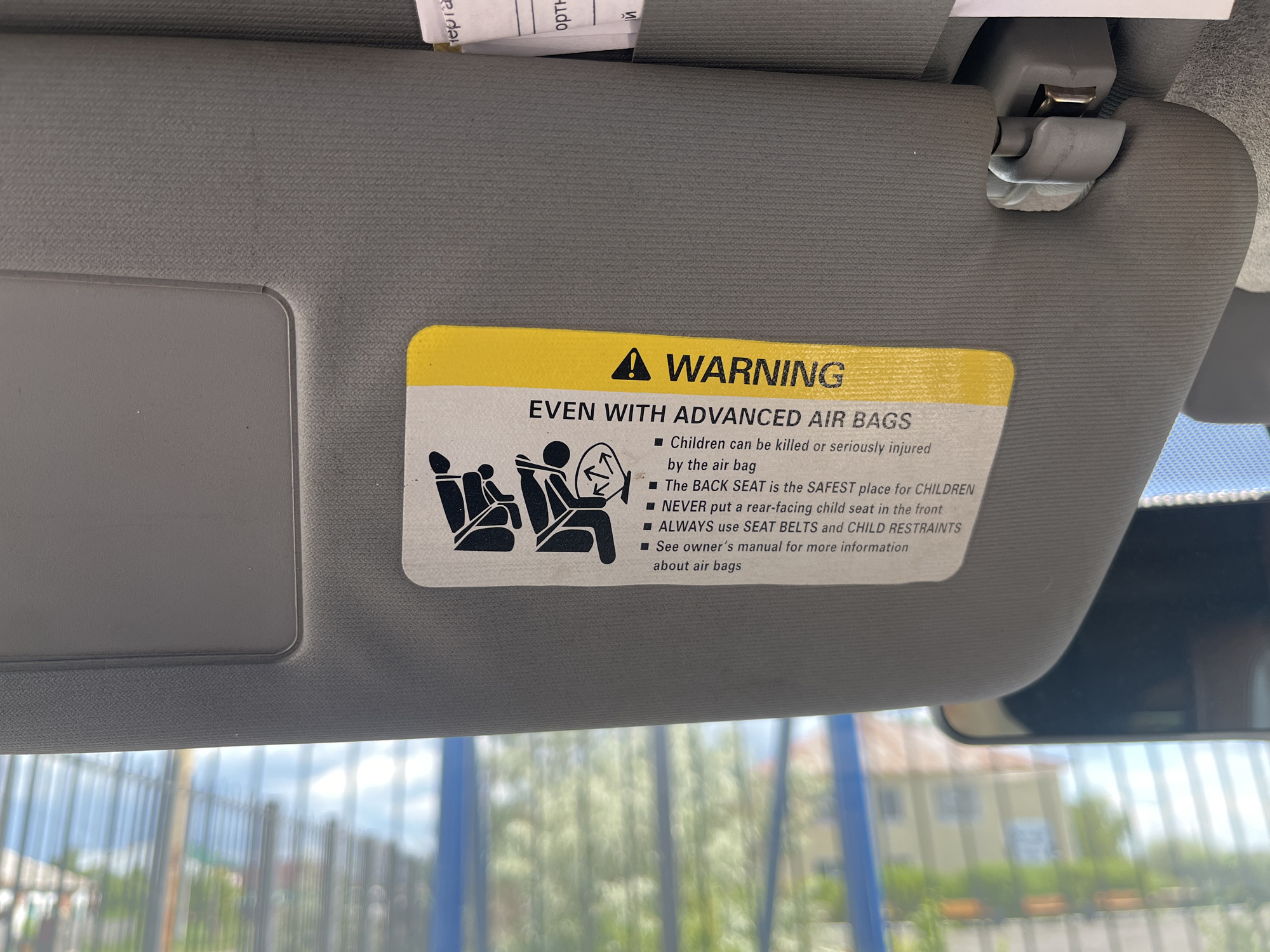 A close-up, eye-level, interior shot captures a gray sun visor in a vehicle, likely in Aqköl, Kazakhstan. Attached to the underside of the sun visor is a yellow warning sticker with black text and graphics. The sticker reads "WARNING" at the top in bold letters, followed by "EVEN WITH ADVANCED AIR BAGS." Below this, there are bullet points and an illustration of child safety seats. The illustration depicts two child safety seats, one rear-facing and one forward-facing, with arrows indicating the direction of impact from an airbag. The bullet points warn about the dangers of airbags to children and recommend placing them in the back seat. The sun visor itself appears to be made of textured fabric and is connected to the car's interior by a plastic clip. The background, visible through the car's windshield, shows a blurred scene of trees, a fence, and buildings, suggesting an outdoor environment.