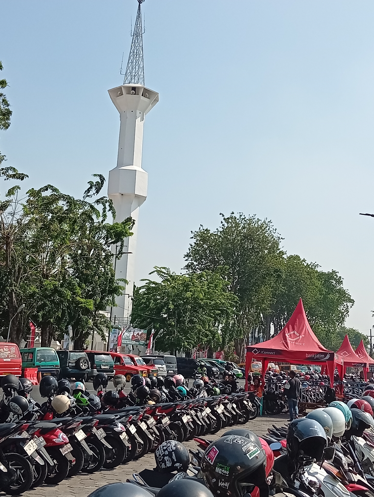 A view of a white minaret tower, topped with a spire and antenna array, rises against a clear blue sky in Sidoarjo, Indonesia. In the foreground, rows of parked motorcycles are visible, many with helmets placed on their seats. Behind the motorcycles, several red tents with "JConnect" and "bankjatim" branding are set up, suggesting an event or promotional activity. Several trees with lush green foliage line the background, with more vehicles, including cars and trucks, parked further back. The scene appears to be a public space on a sunny day.