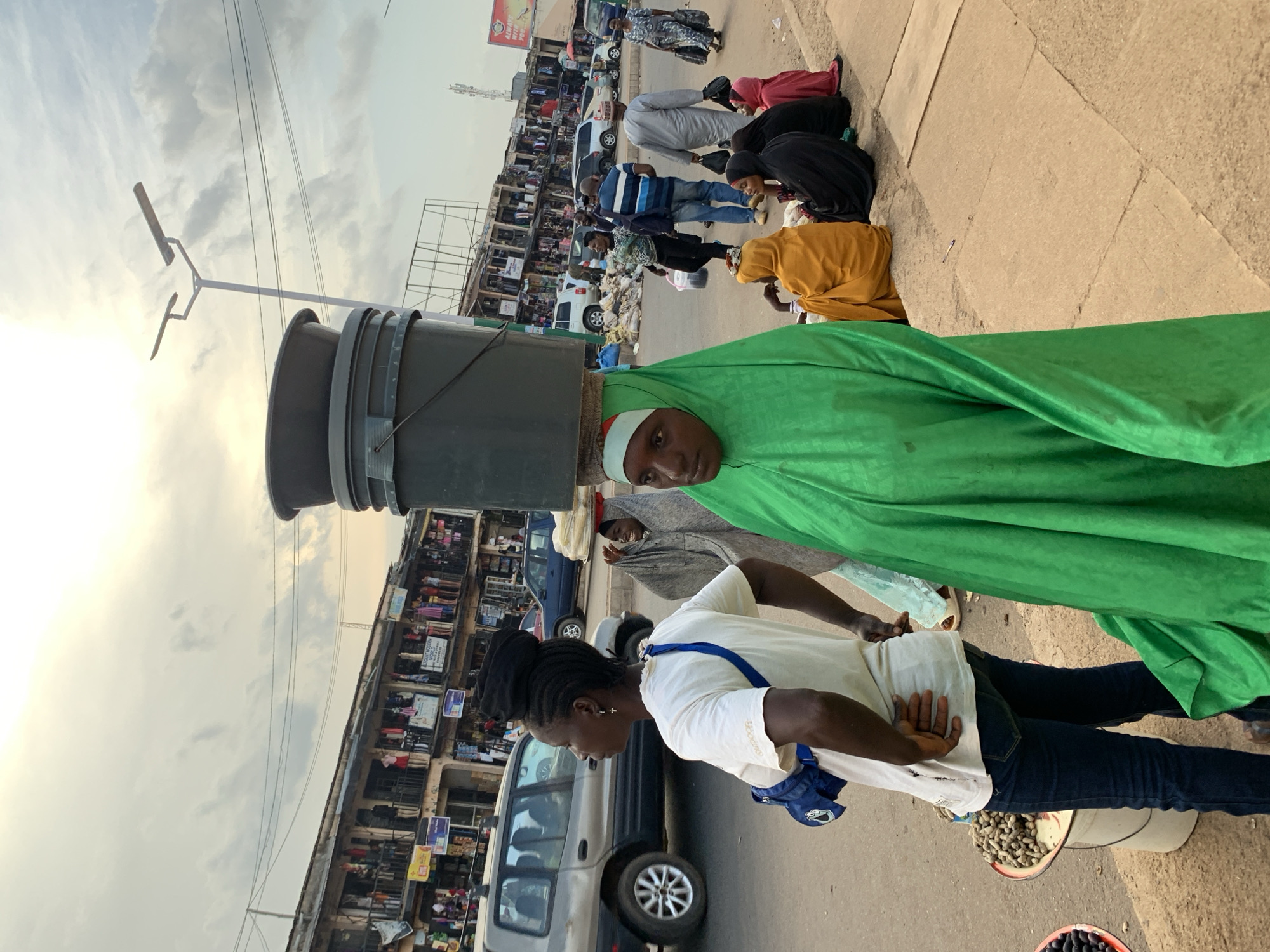 The image depicts a street scene in Jos, Nigeria, with a focus on a woman carrying a large bucket on her head, draped in a vibrant green garment. She is positioned in the foreground, with her head turned slightly to the side, revealing a portion of her face. Behind her, several other individuals are visible, some sitting on the ground along the edge of a paved walkway. One person wears a bright yellow headscarf, another a black headscarf, and a third a burgundy garment. Further in the background, a row of market stalls or shops lines the street, with various goods displayed. Cars are parked or driving on the road adjacent to the market.
The time of day appears to be late afternoon or early evening, suggested by the warm, diffuse light and the long shadows cast by the overhead structures and people. The sky is cloudy, indicating potentially overcast weather. The presence of a street light pole suggests an urban environment. The overall atmosphere seems busy, typical of a market area. There is no visible text that is clearly legible in the image. The people's postures and clothing suggest everyday life and commerce in this Nigerian city.