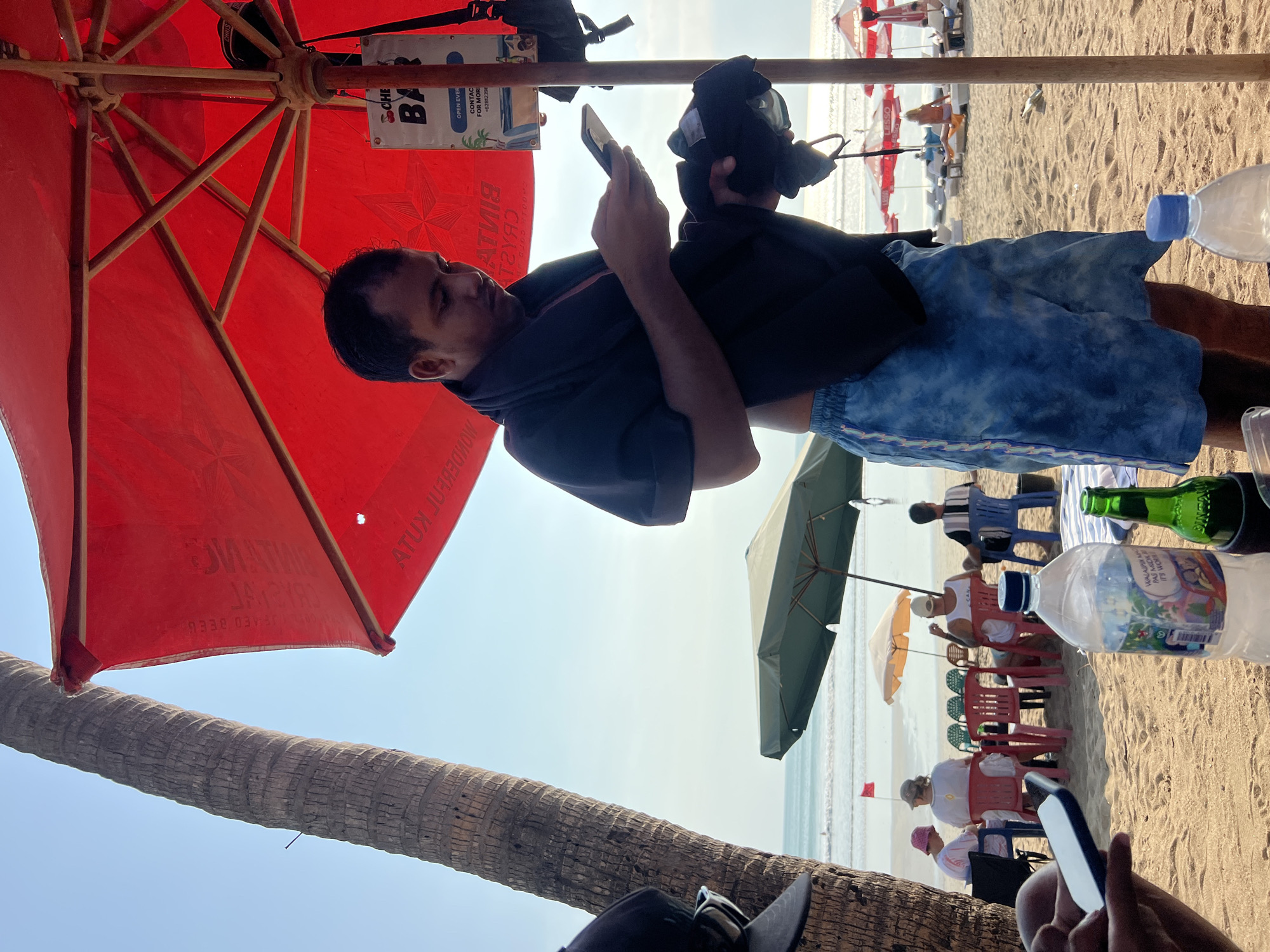 A man stands on a sandy beach in Kuta, Indonesia, under a large red umbrella. He is wearing blue tie-dye shorts and has a black towel draped over his shoulders. He is looking down at his phone, holding another dark-colored fabric in his left hand. A partially visible sign with "BAR" written on it is attached to the umbrella pole. In the background, the ocean can be seen with gentle waves. Several beach chairs and umbrellas are set up on the sand, with a few people seated in them. A red flag is planted in the sand near the water. A large palm tree trunk is visible on the left side of the frame. A plastic water bottle and a green bottle are on the sand in the foreground.